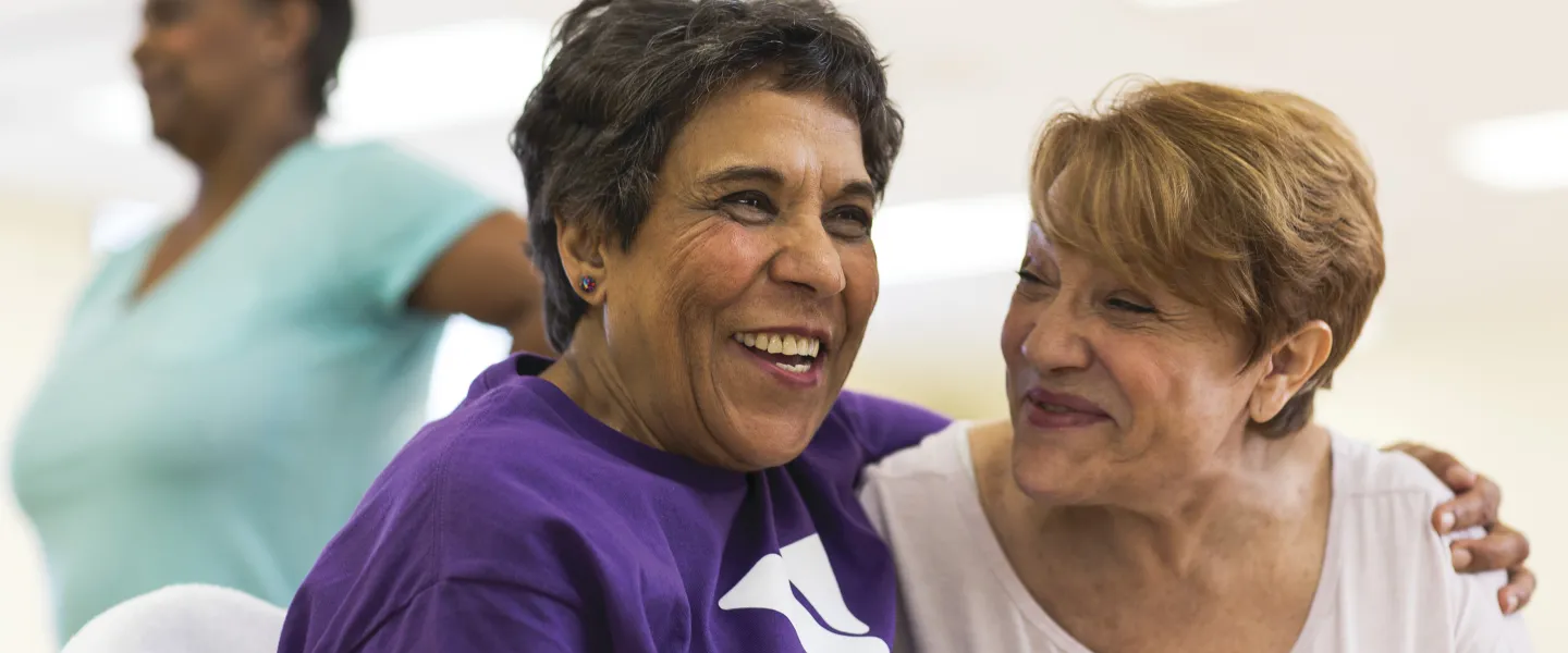 Two smiling women with arms around each other, one in purple shirt with logo, another woman in the background.