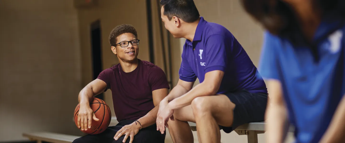 Two men and a blurred person sit on a bench in a gymnasium. One man holds a basketball. They appear to be talking during a break.