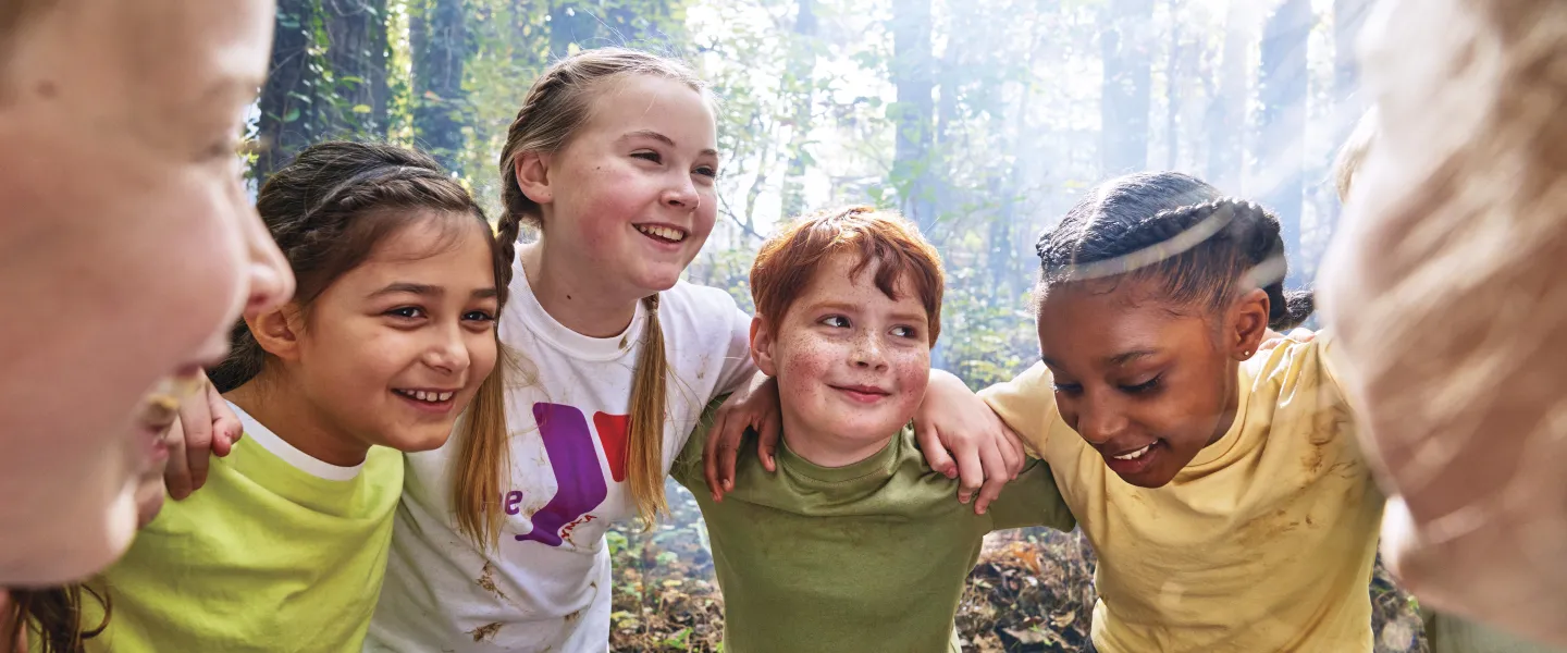 Group of kids huddled together, smiling, with arms around each other in a forest setting, enjoying camp activities.