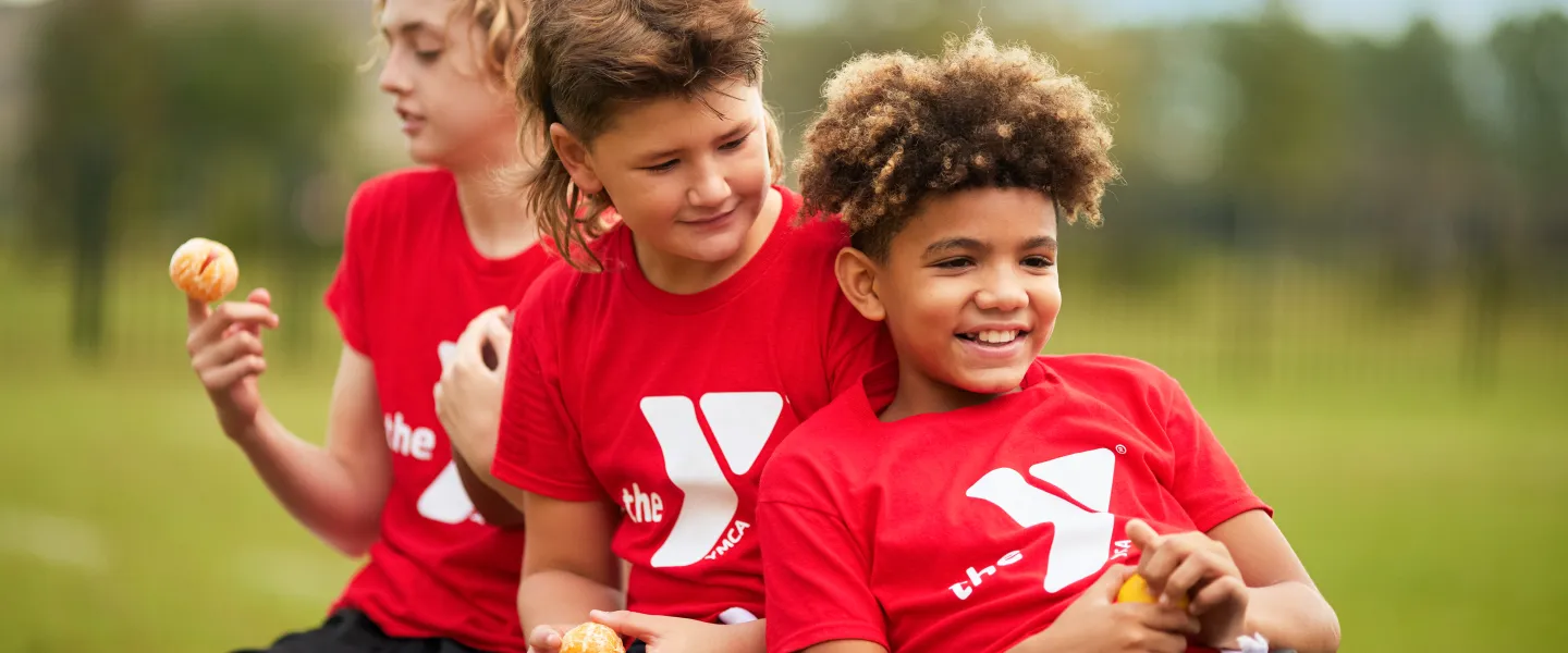 Three young children in red shirts with white logos and black shorts smile, holding small balls while sitting outside on a bench.