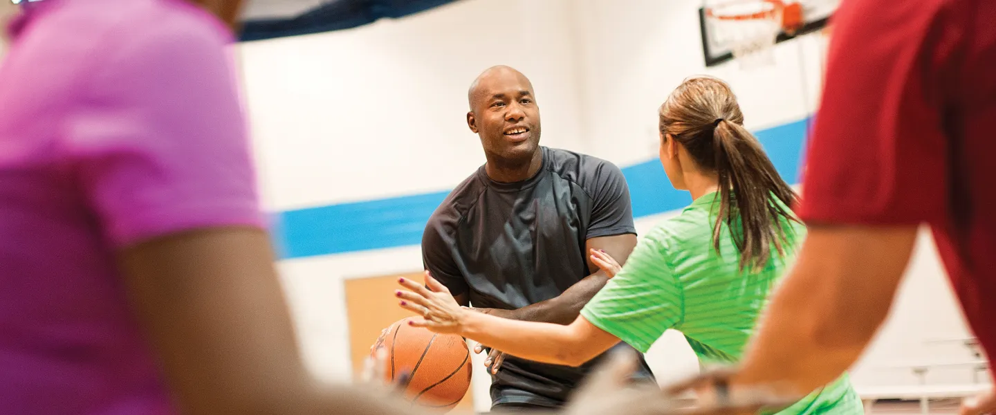 Adults playing basketball in a gymnasium. One man smiles and passes the ball to a woman wearing green. Several other people are in the scene.