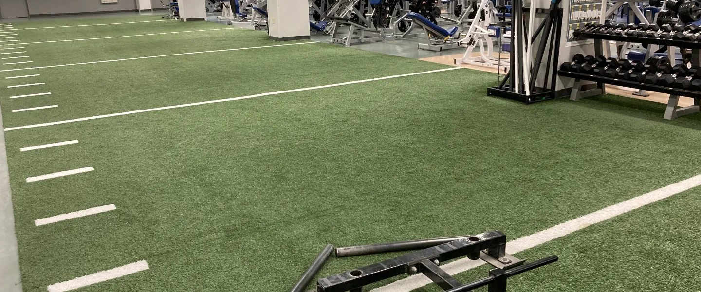 Indoor turf field with fitness equipment in the background, including weight machines and free weights; a strength training center.
