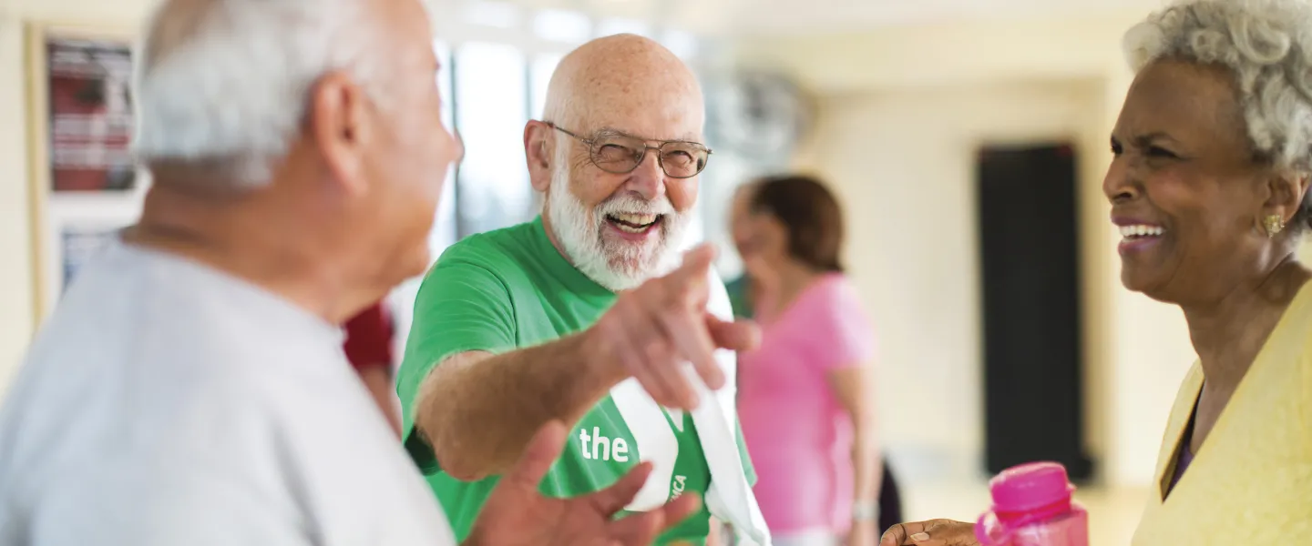 Senior adults laughing and chatting after a fitness class. A man in green points, others hold water bottles. Light-filled room.
