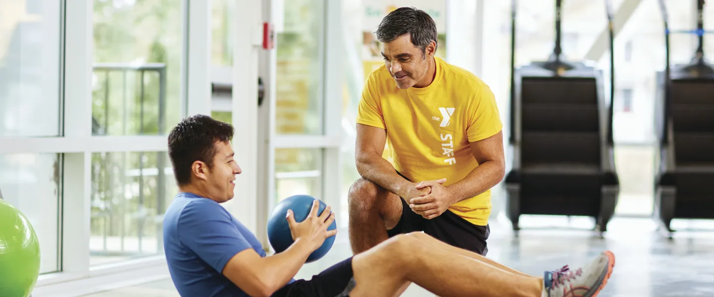 Man doing core workout with a medicine ball while a trainer kneels nearby, watching the exercise.