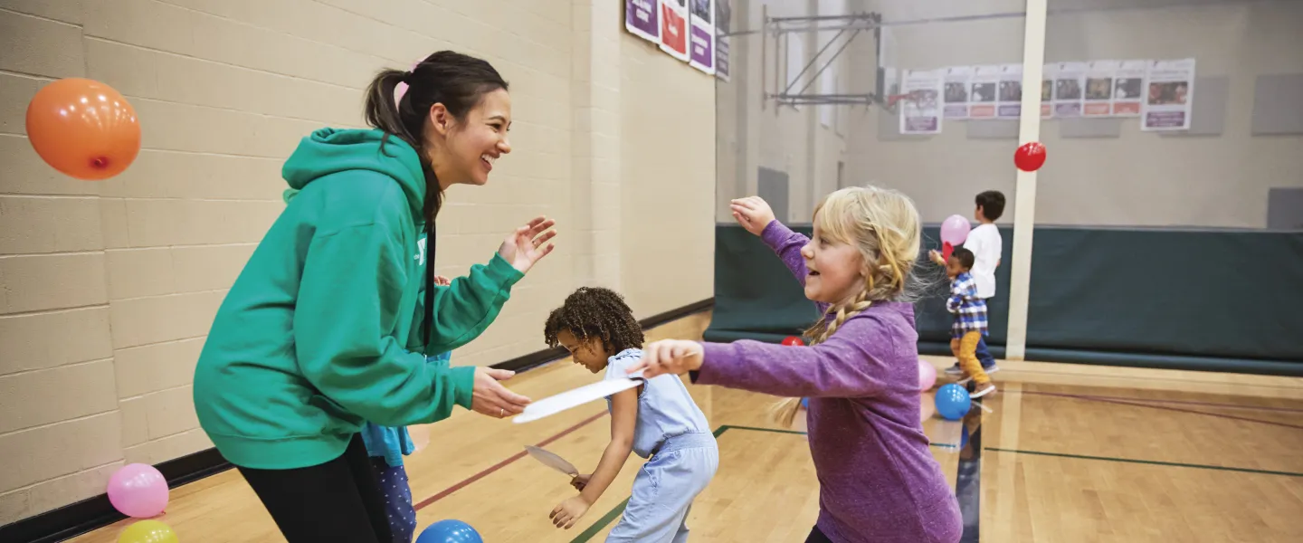 Children playing games in a gymnasium, with an instructor holding a paper strip. Kids enjoy active recreation, smiling and laughing. Colorful balloons scattered on the wooden floor.