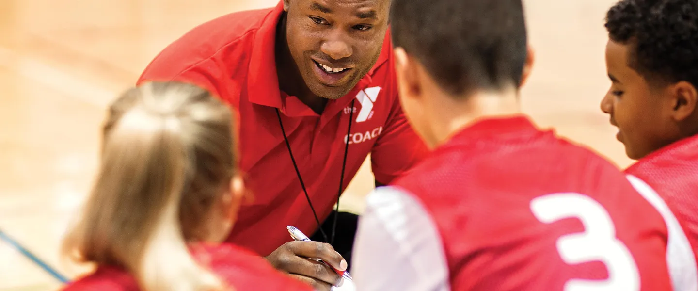 A coach in a red shirt with "COACH" talks to kids in red team shirts numbered one and three on a basketball court.