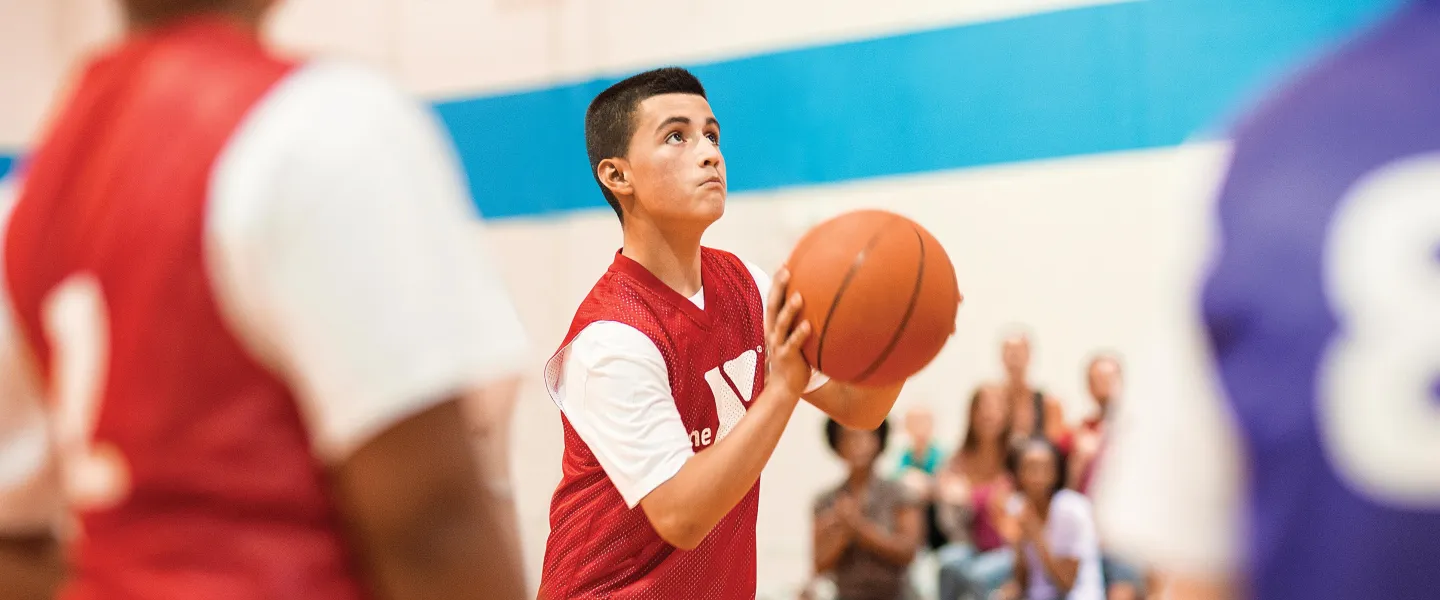 Teenager in red jersey shoots basketball in gymnasium, looking up towards the hoop. Spectators and other players blurred in background.