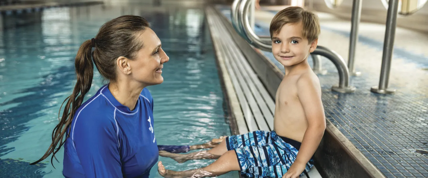 A young boy in swim trunks sits poolside, smiling at a female instructor partially submerged in the water.  They appear to be participating in a swim lesson.