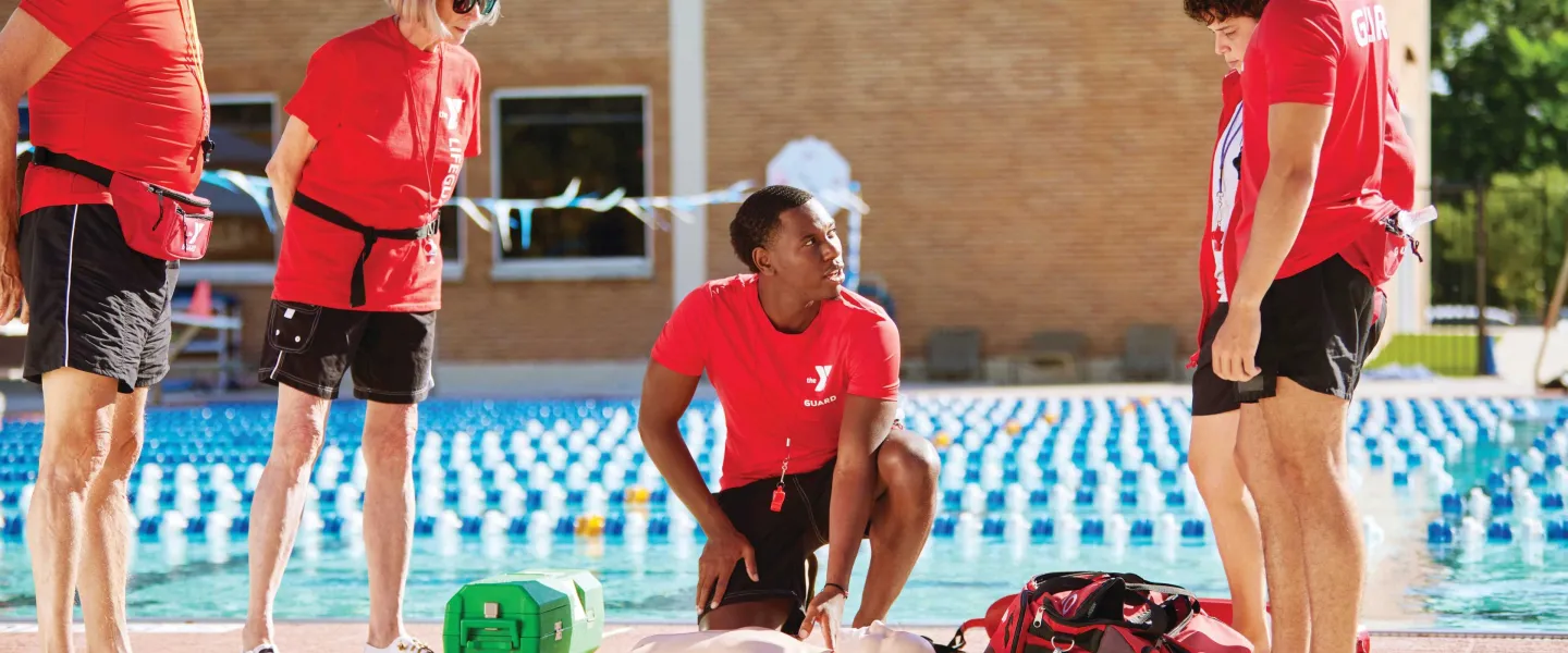 Lifeguards in red shirts practice CPR on a training dummy by the pool with others watching. A green first aid kit and a red bag lay nearby.