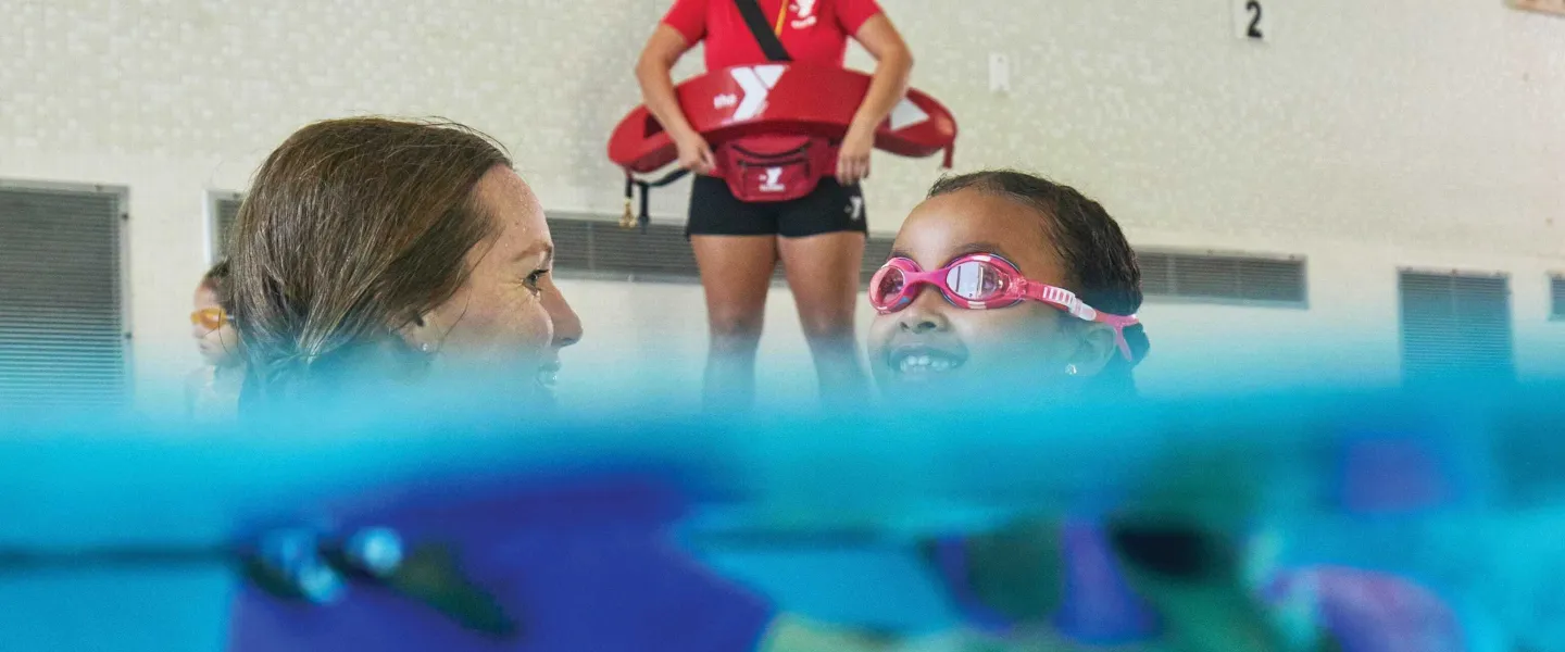 Smiling child in pink goggles learns to swim with an instructor; lifeguard watches over them at indoor pool, safety first.