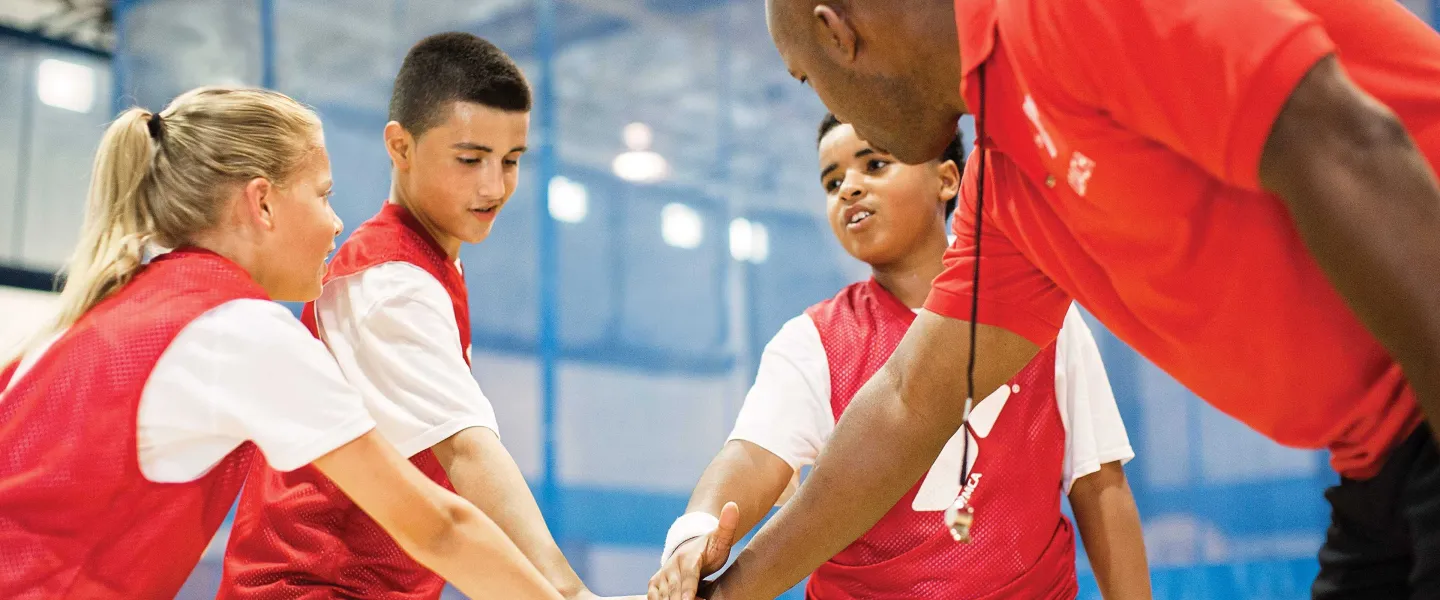 A coach and three kids in red vests with hands stacked in the middle in a team spirit gesture, indoors.
