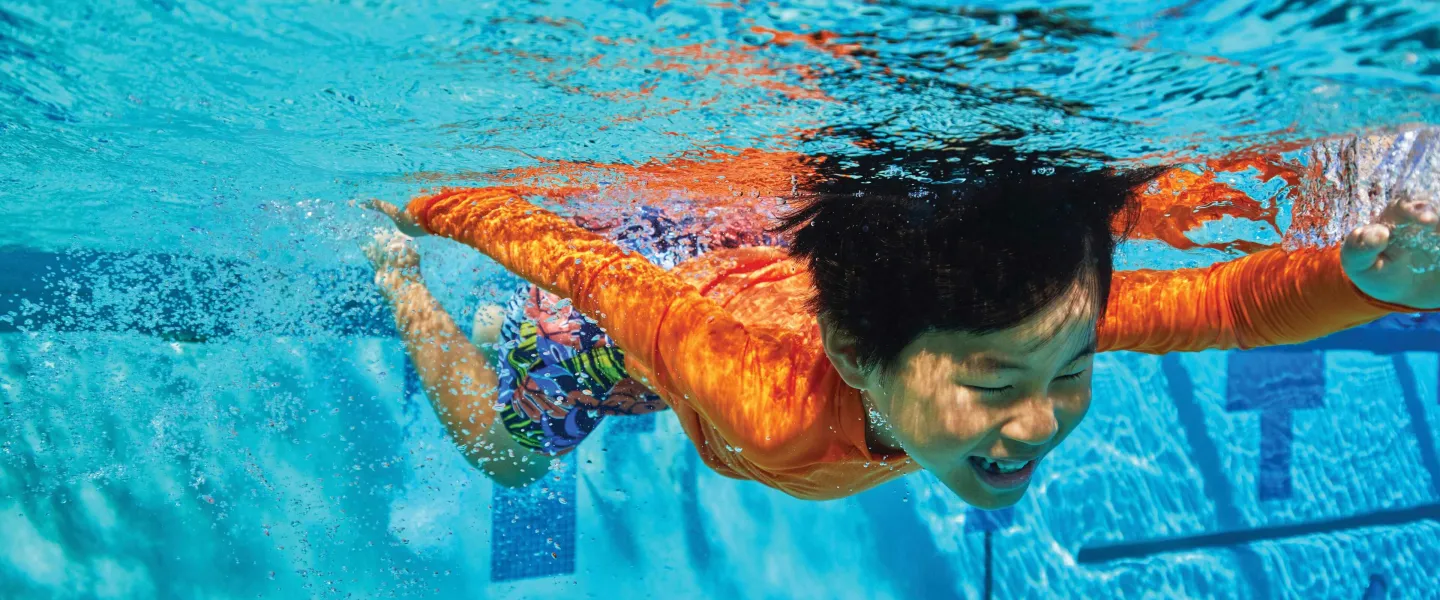 Young boy in an orange shirt swims underwater in a pool, arms stretched forward and smiling. Clear blue water surrounds him.