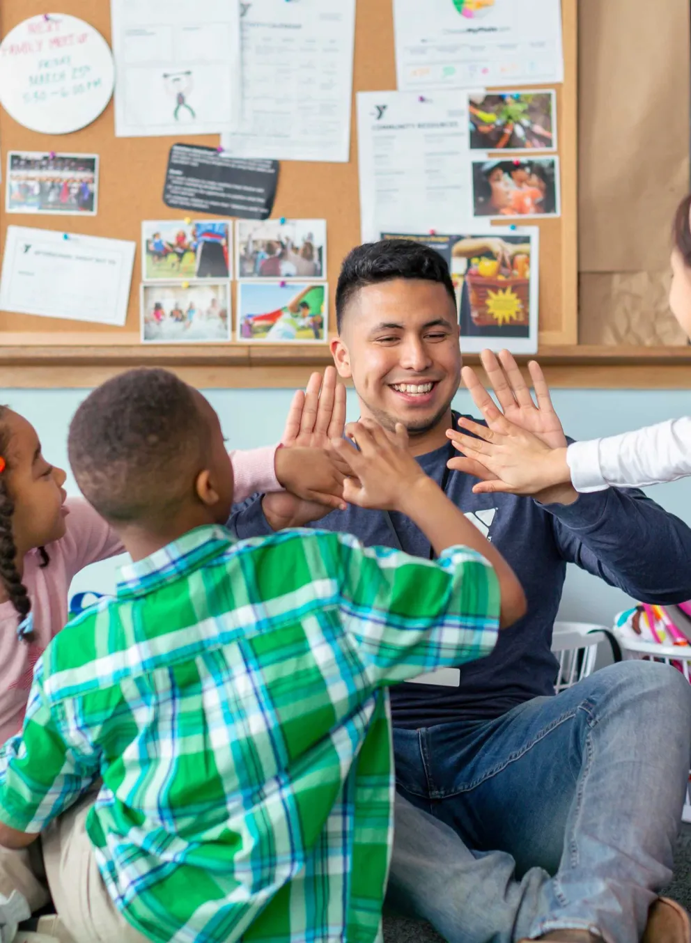 A counselor and four children in a circle sit on the floor, all giving high-fives in a playful classroom scene.