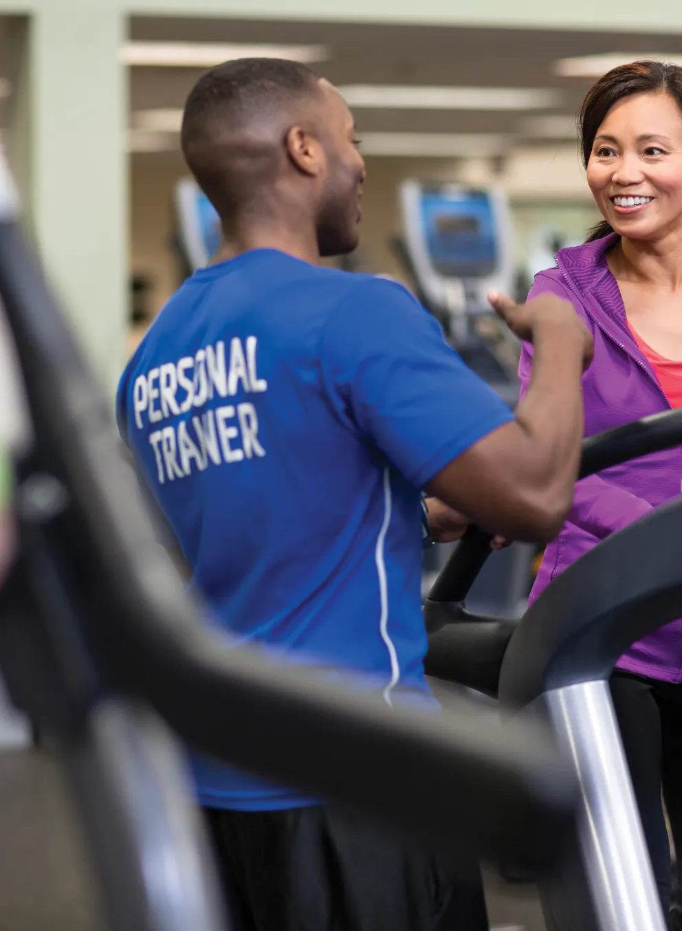 Personal trainer in blue shirt talking with a woman on treadmill; wellness center workout is ongoing. Another person is running on a machine.