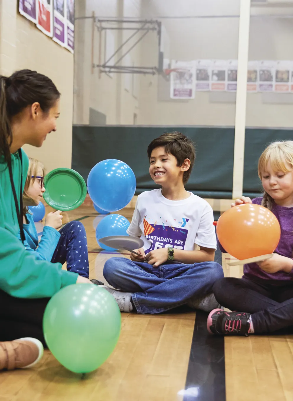 Children and an instructor are seated in a gymnasium, holding colorful balloons and toys; they smile and appear happy. The instructor in a green hoodie is interacting with kids.