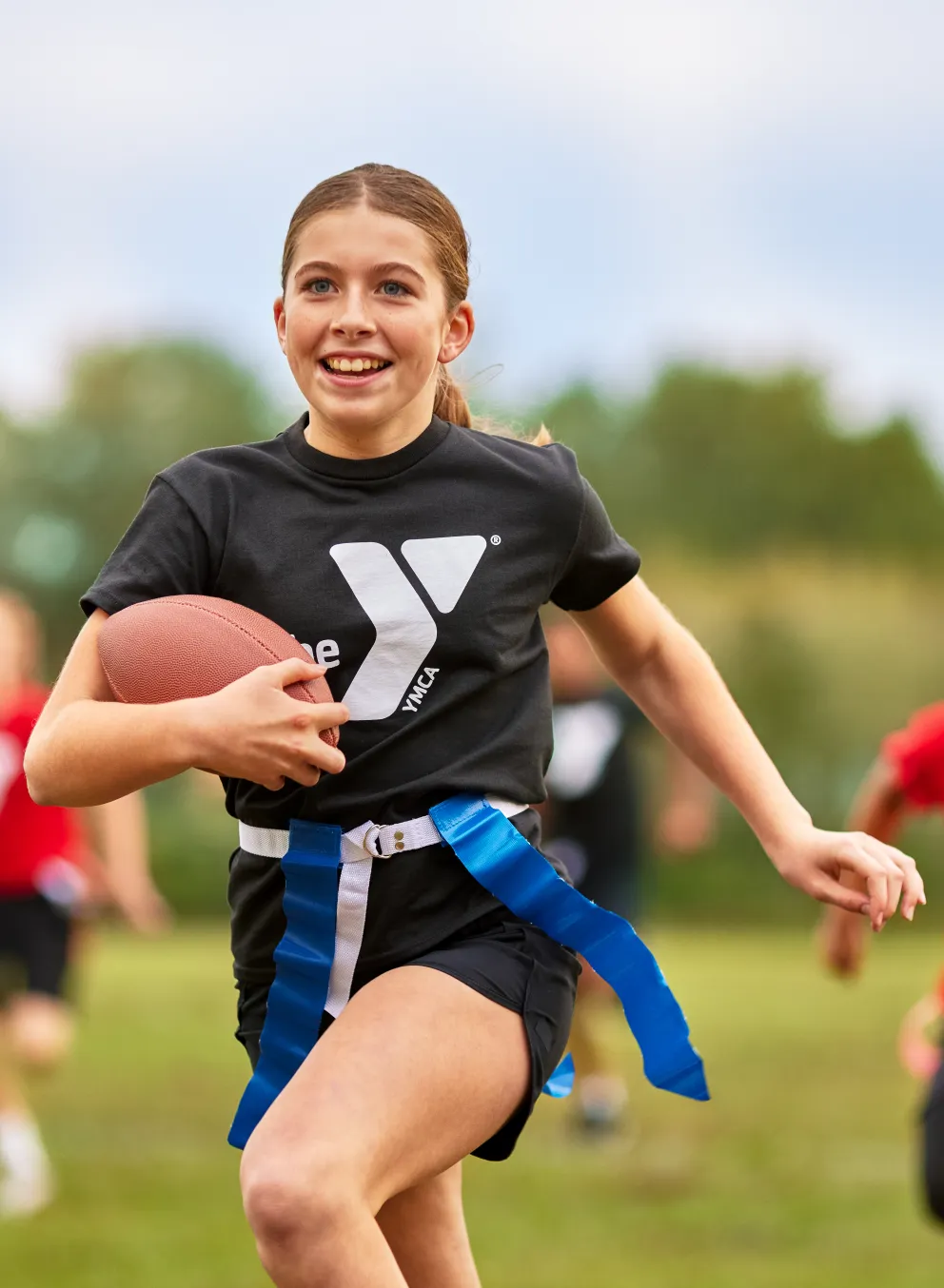 A girl with a football runs on a green field, with flags attached to her waist, smiling. Two children in red shirts run behind her.