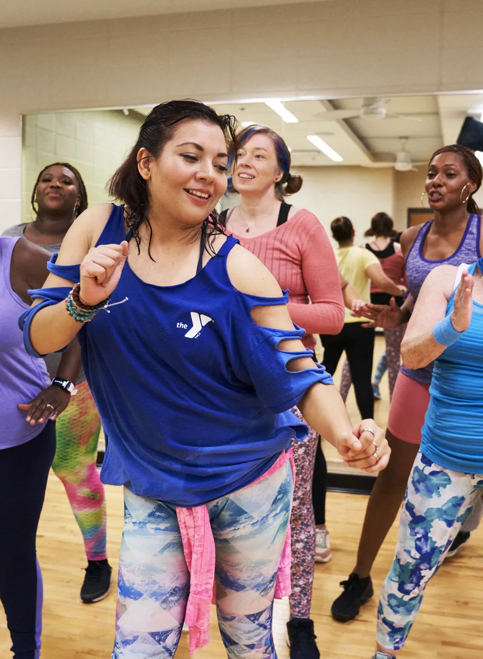Group of people enjoying fitness class. Women and men dancing in brightly lit studio. One woman in blue top is clapping and smiling.