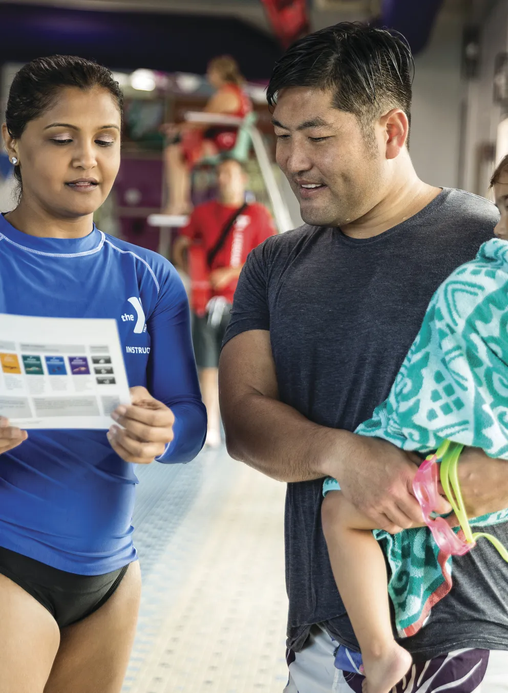 Instructor in blue shirt holding a program paper discussing with a father, holding a toddler wrapped in a towel, by the indoor pool.