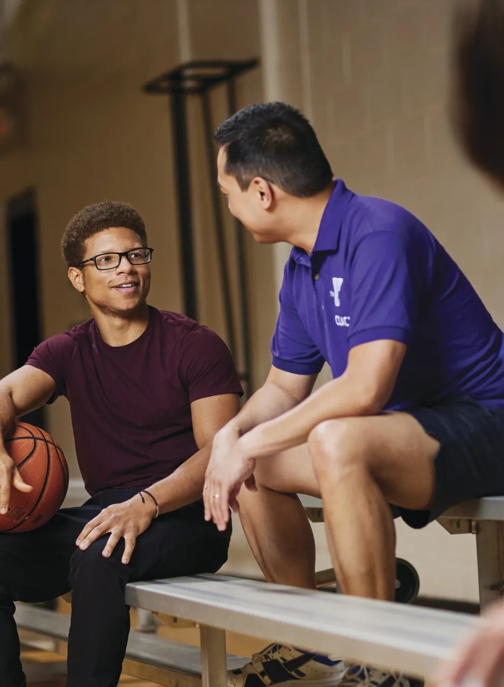 Two men and a blurred person sit on a bench in a gymnasium. One man holds a basketball. They appear to be talking during a break.