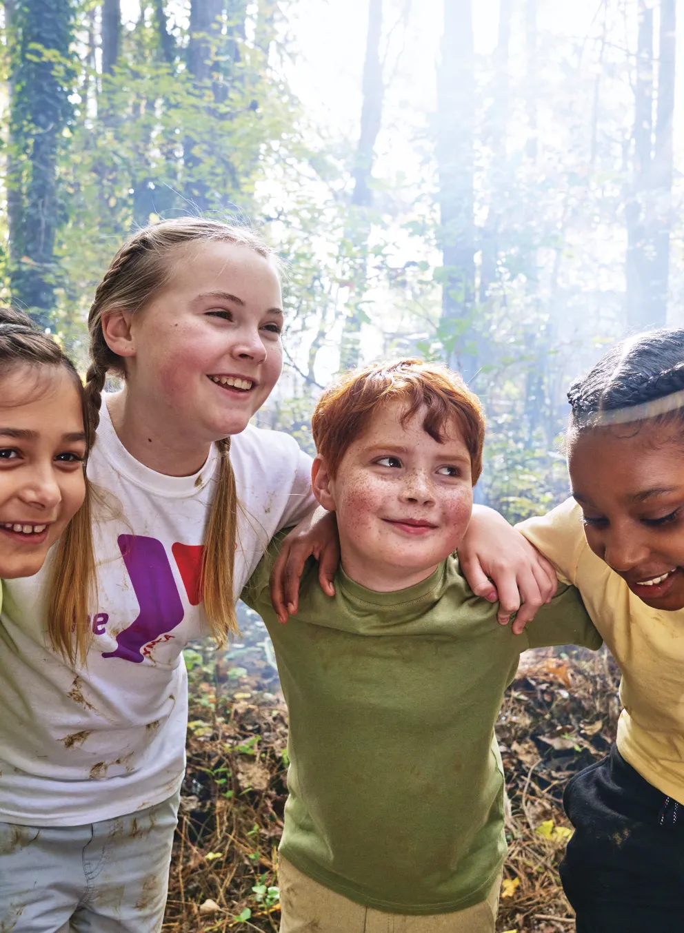 Group of kids huddled together, smiling, with arms around each other in a forest setting, enjoying camp activities.