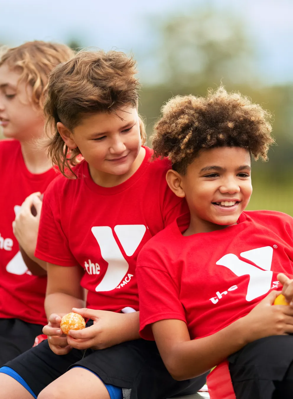 Three young children in red shirts with white logos and black shorts smile, holding small balls while sitting outside on a bench.