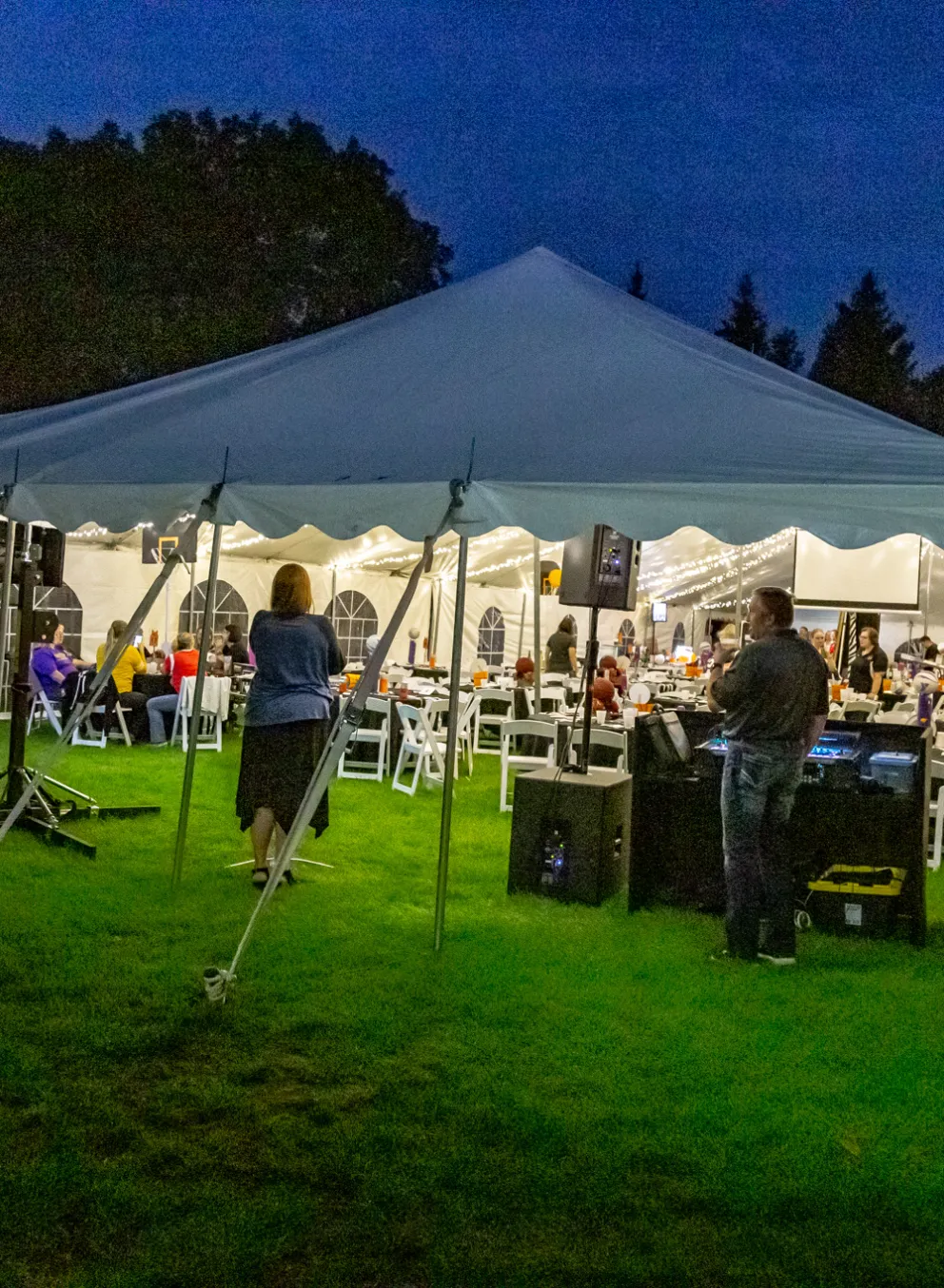 Large white tent set on green grass, tables and people inside, a DJ playing music for a community event at night.