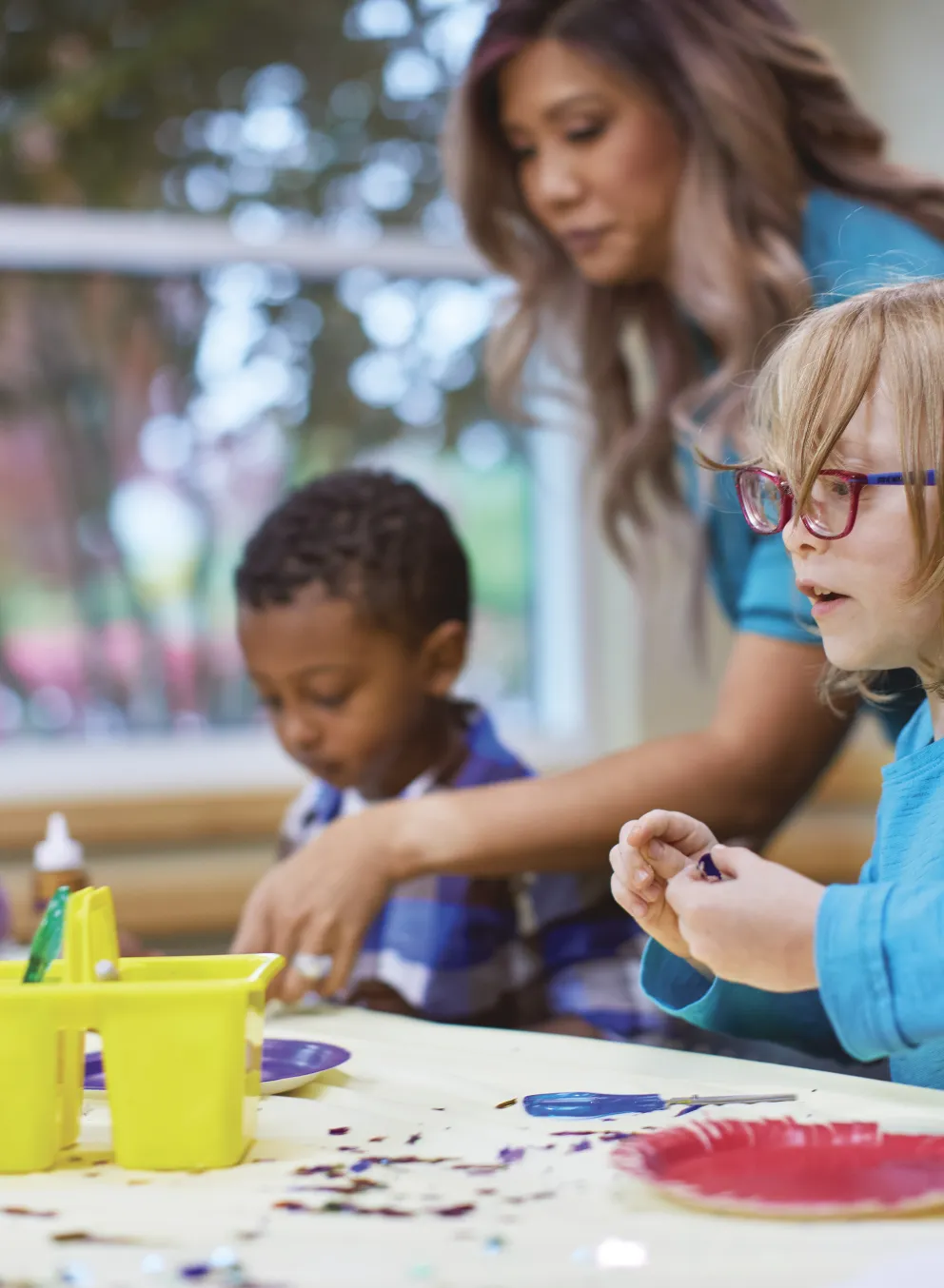 Children in childcare program gather around a table. An adult supervises as the kids work on crafts with paint and paper plates.