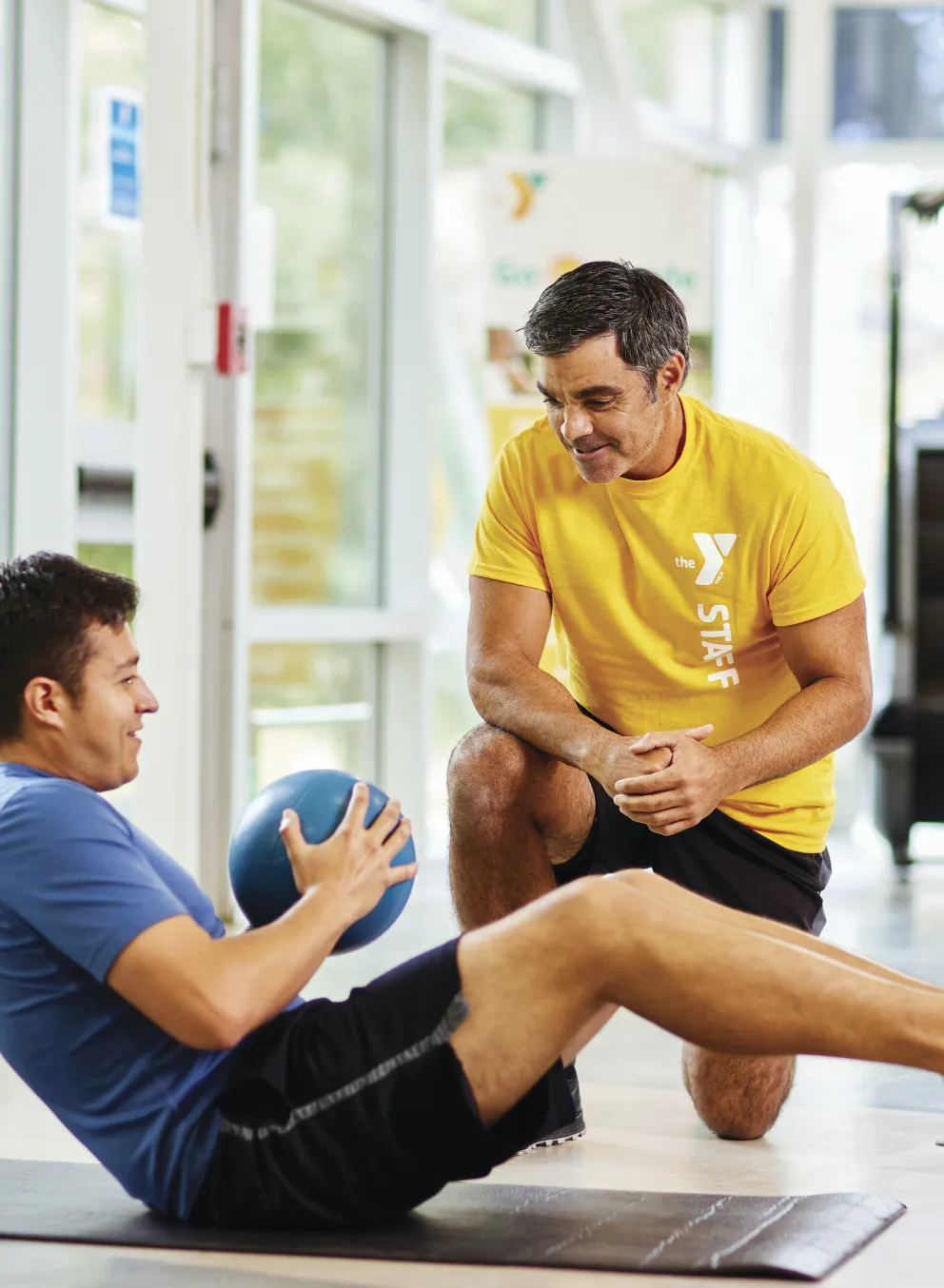 Man doing core workout with a medicine ball while a trainer kneels nearby, watching the exercise.