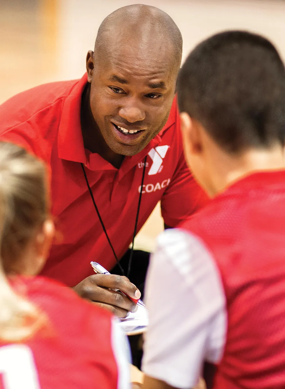 A coach in a red shirt with "COACH" talks to kids in red team shirts numbered one and three on a basketball court.