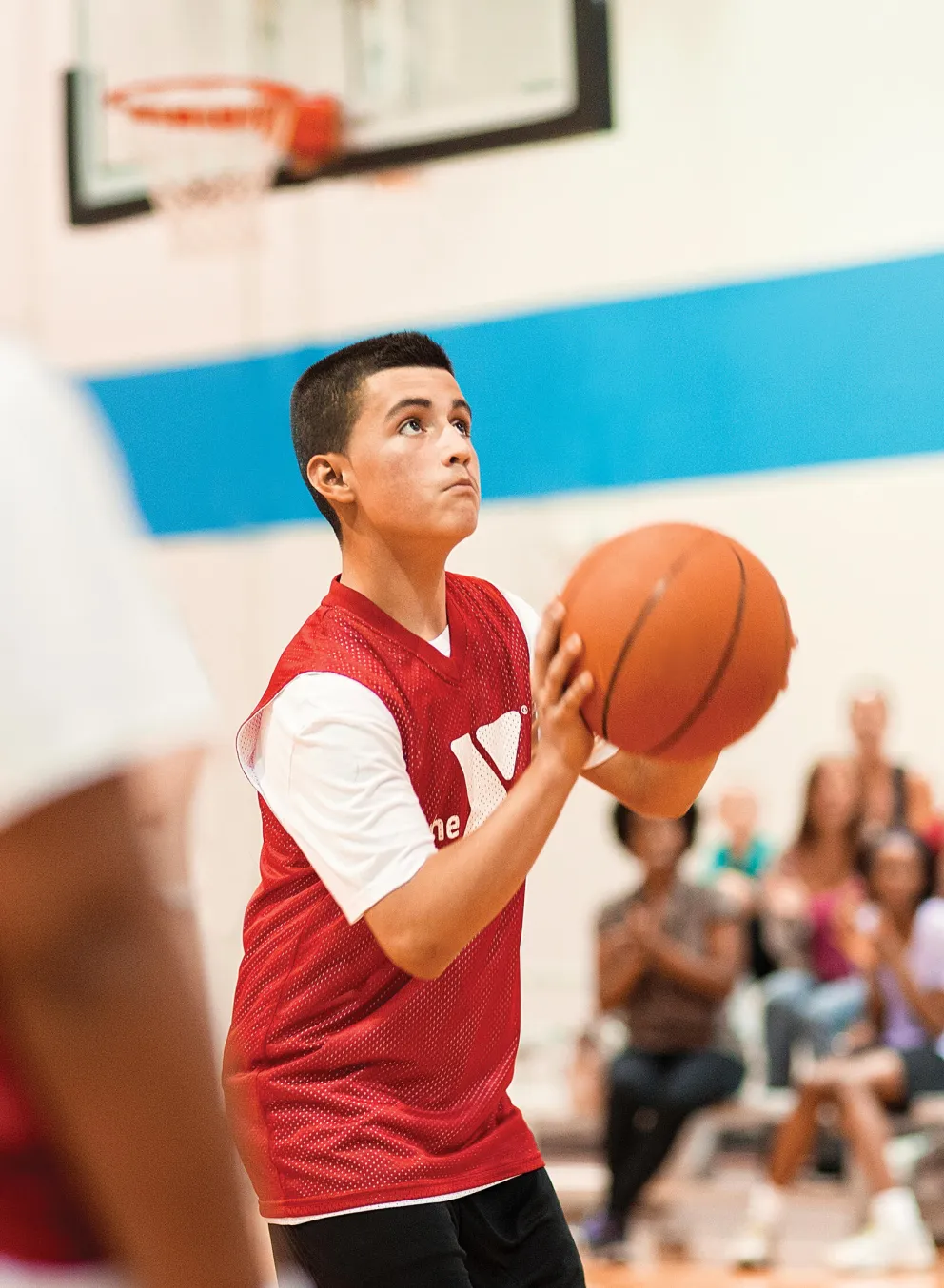 Teenager in red jersey shoots basketball in gymnasium, looking up towards the hoop. Spectators and other players blurred in background.