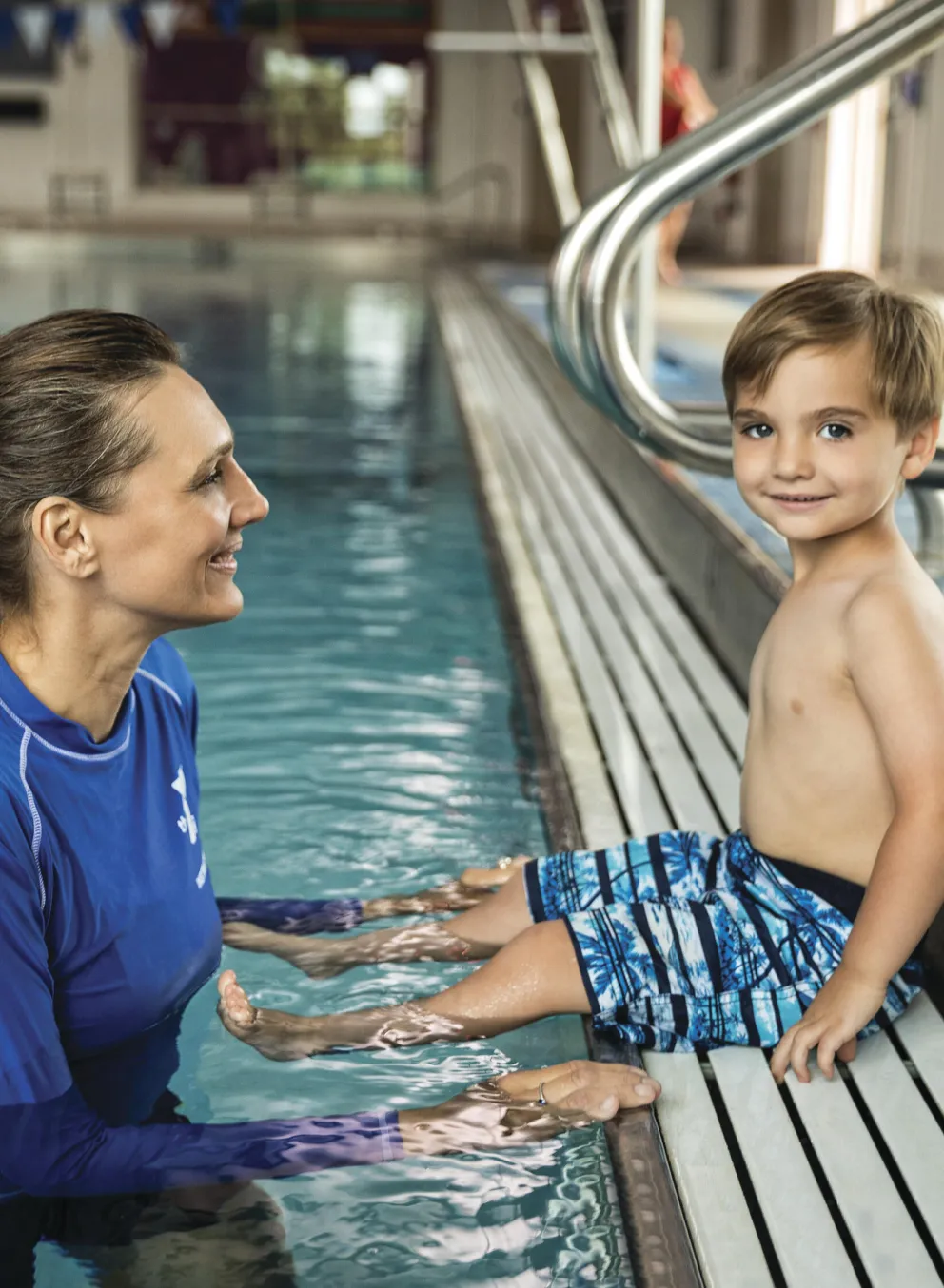 A young boy in swim trunks sits poolside, smiling at a female instructor partially submerged in the water.  They appear to be participating in a swim lesson.