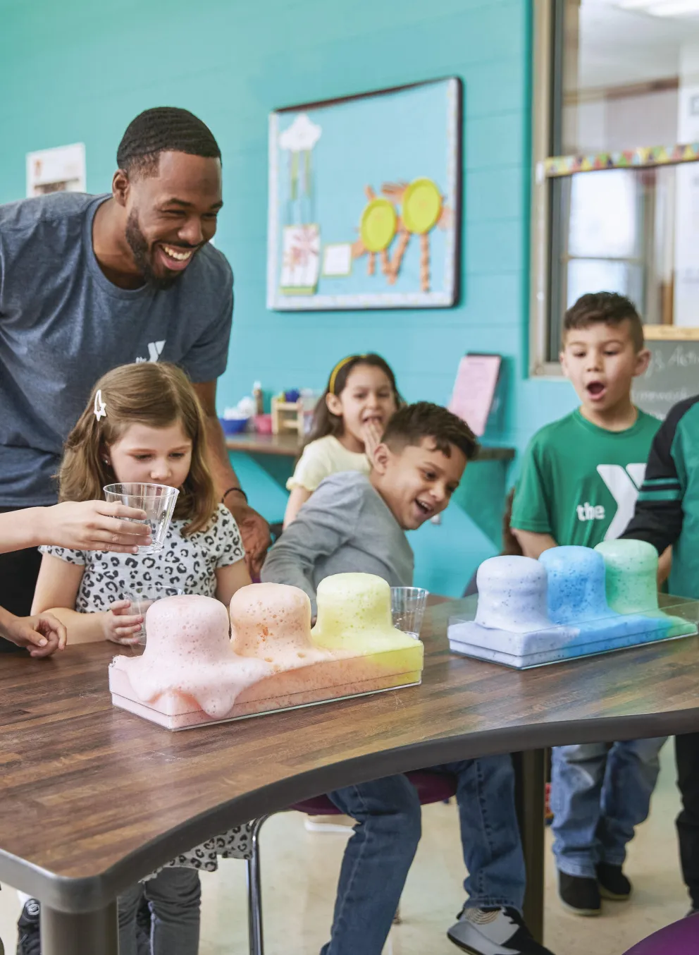 Kids conduct a science experiment with colorful substances, guided by a smiling instructor.  They're engaged and excited, working together at a table in a brightly colored classroom.