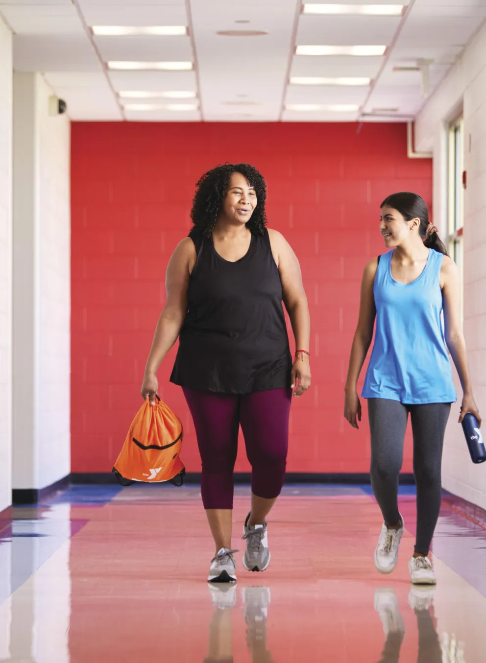 Two women stroll down a hallway, chatting; one carries an orange gym bag, the other a water bottle.  A third person, wearing a brightly colored shirt, walks ahead.  Colorful posters line the hallway walls.