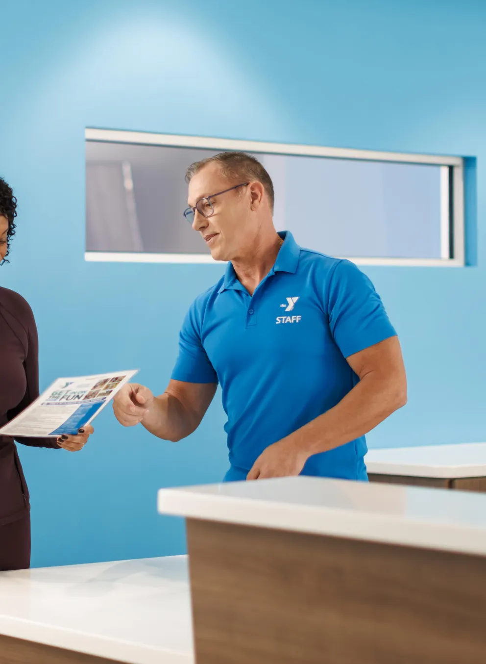 A woman and staff member review a brochure at the front desk.  Happy expressions.  Light blue walls.