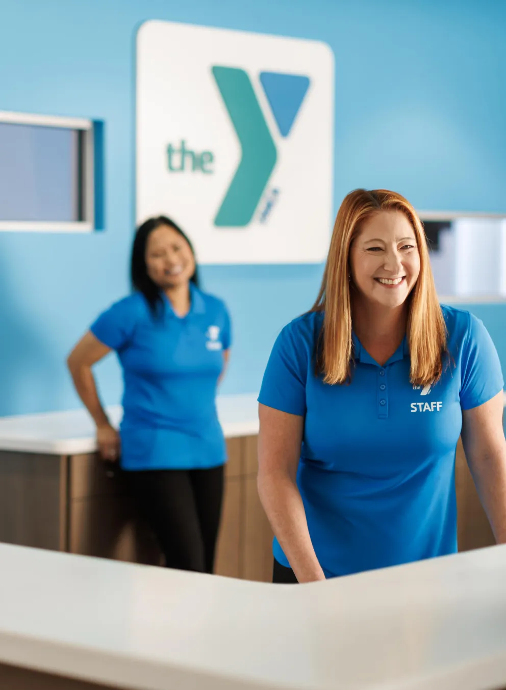 Smiling staff members at a reception desk greet a visitor.  One staff member's shirt says "STAFF".  Happy atmosphere.