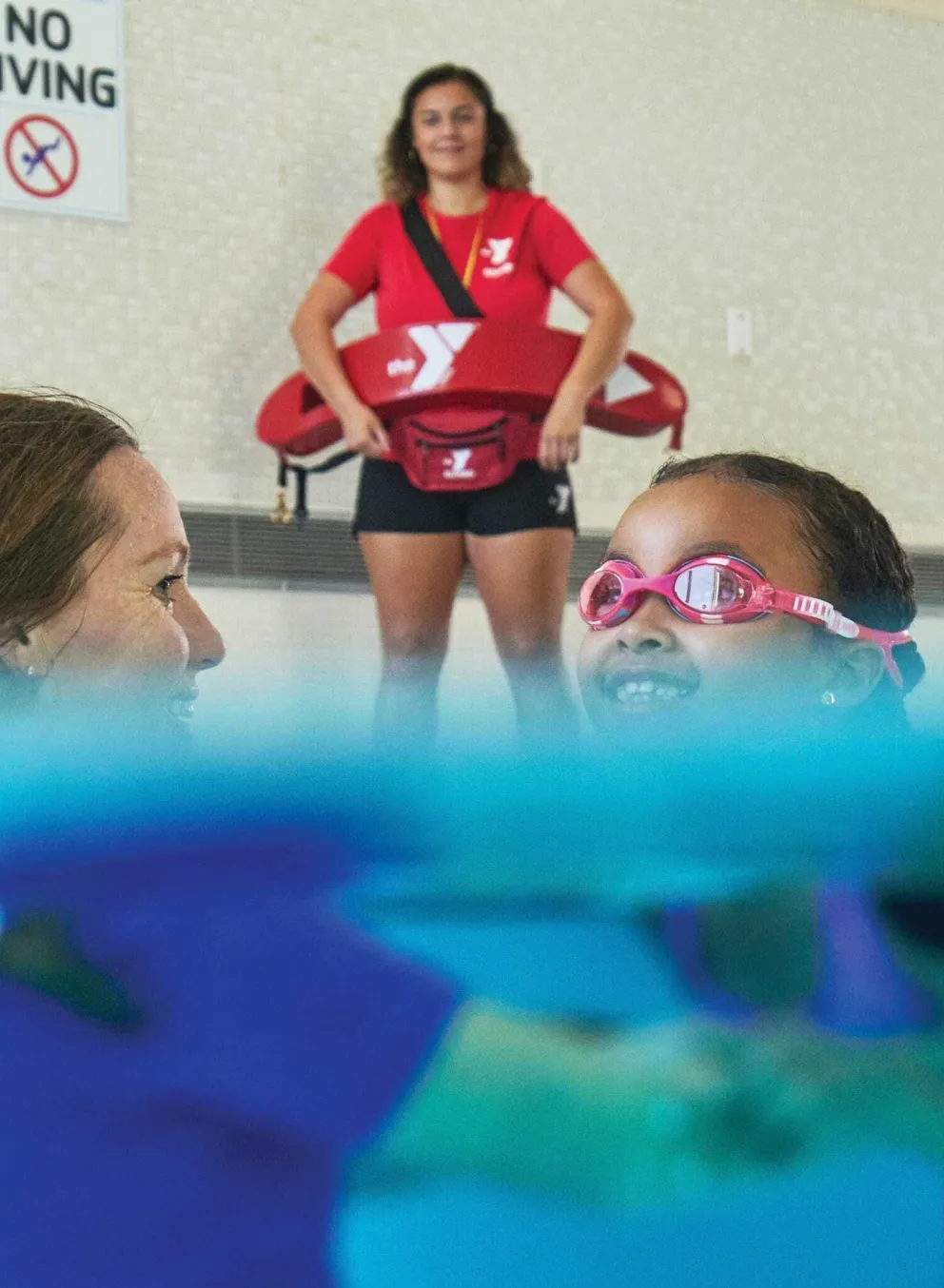 Smiling child in pink goggles learns to swim with an instructor; lifeguard watches over them at indoor pool, safety first.