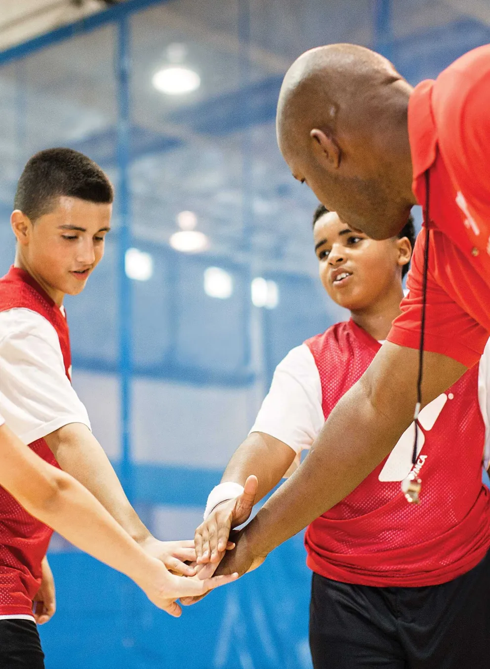 A coach and three kids in red vests with hands stacked in the middle in a team spirit gesture, indoors.