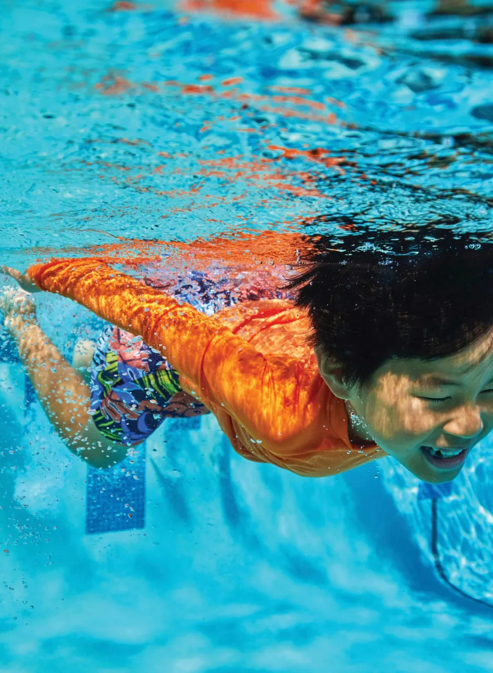 Young boy in an orange shirt swims underwater in a pool, arms stretched forward and smiling. Clear blue water surrounds him.