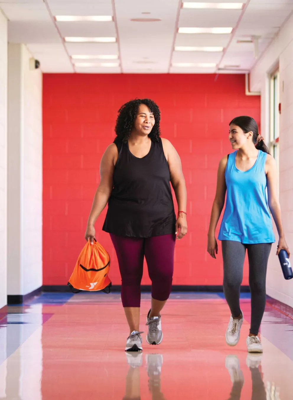Two women are walking and talking in a brightly lit hallway with a third person walking away. One woman carries a bag, another a water bottle.
