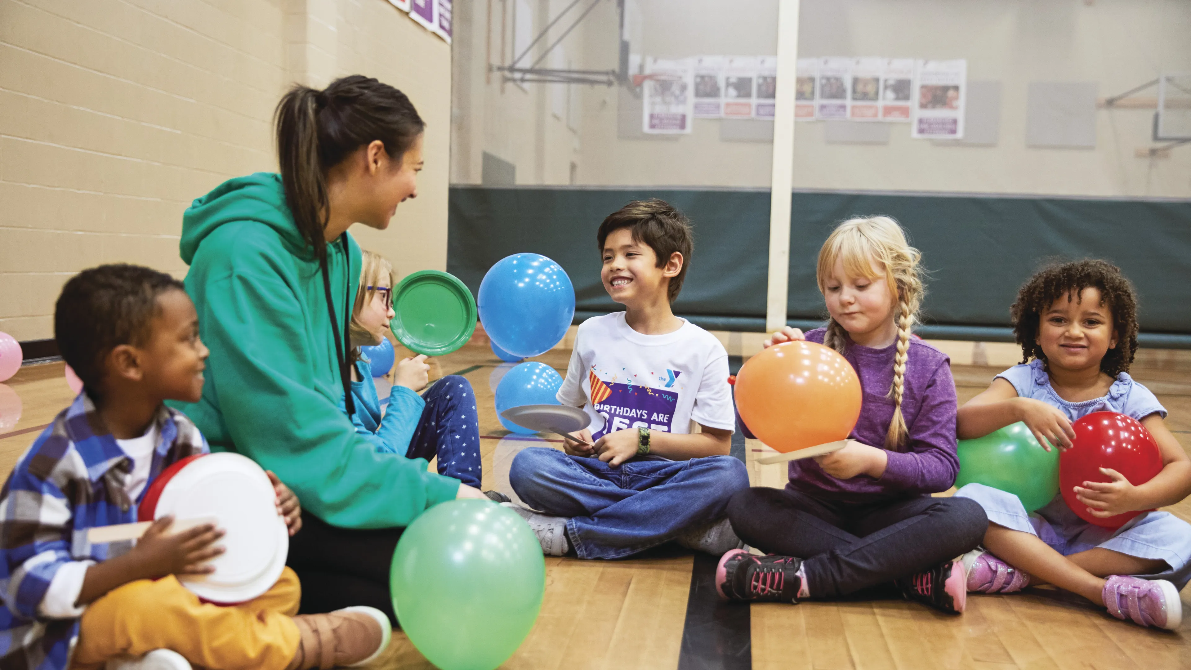 Children and an instructor are seated in a gymnasium, holding colorful balloons and toys; they smile and appear happy. The instructor in a green hoodie is interacting with kids.