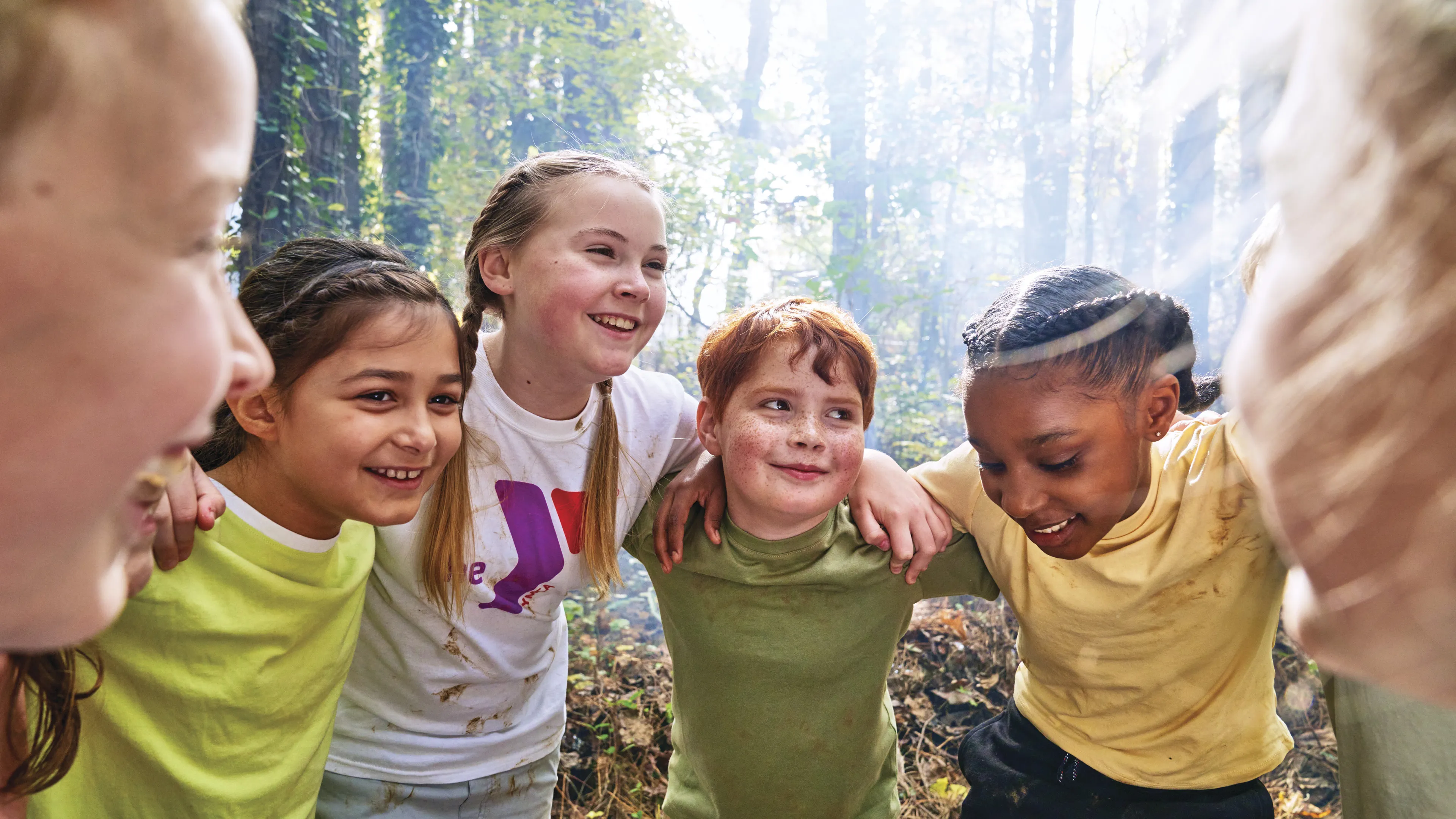Group of kids smiling and hugging outdoors in a forest. Youngsters wearing casual clothes, possibly at a camp, showing joy and friendship.