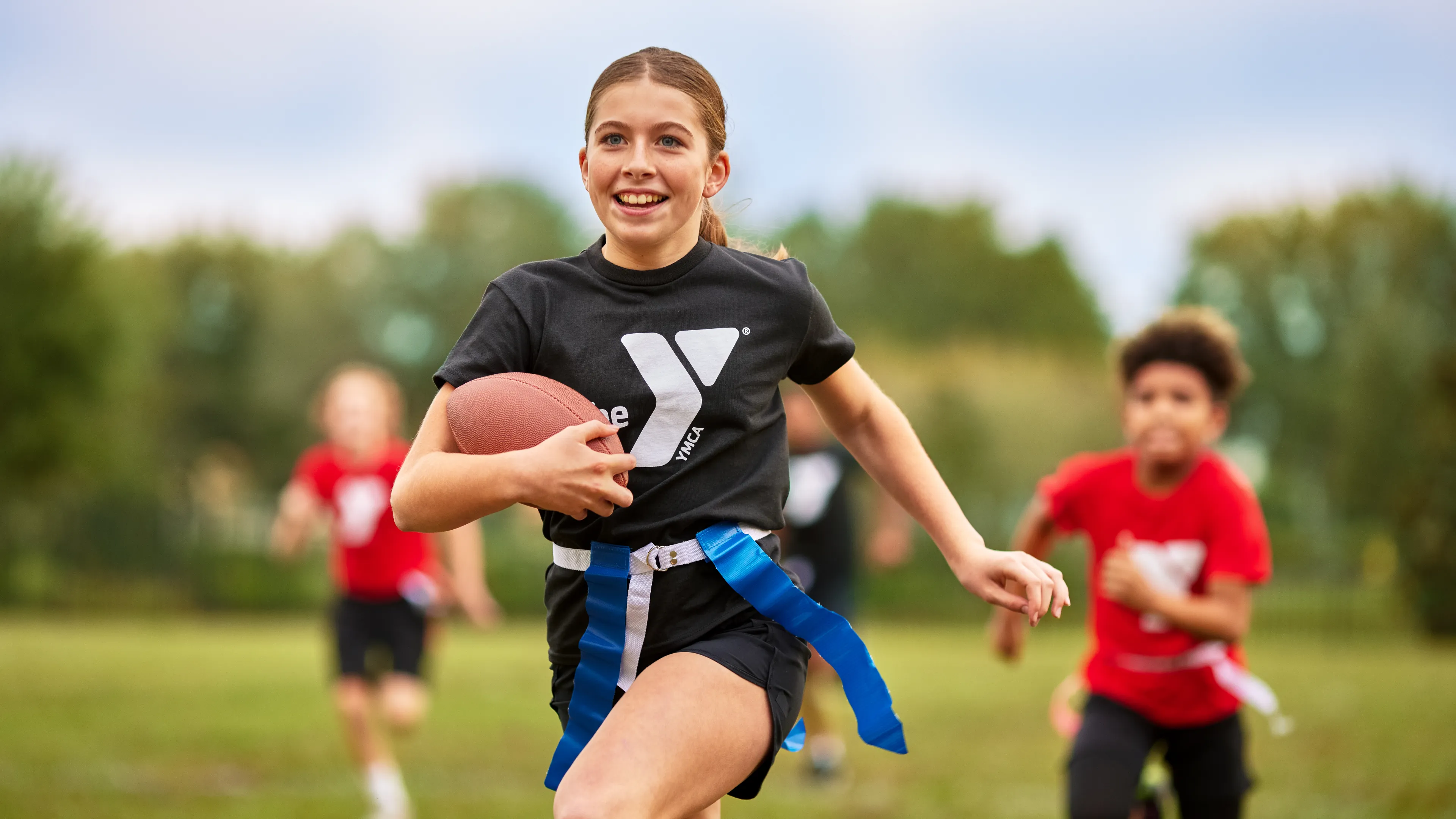 A girl with a football runs on a green field, with flags attached to her waist, smiling. Two children in red shirts run behind her.