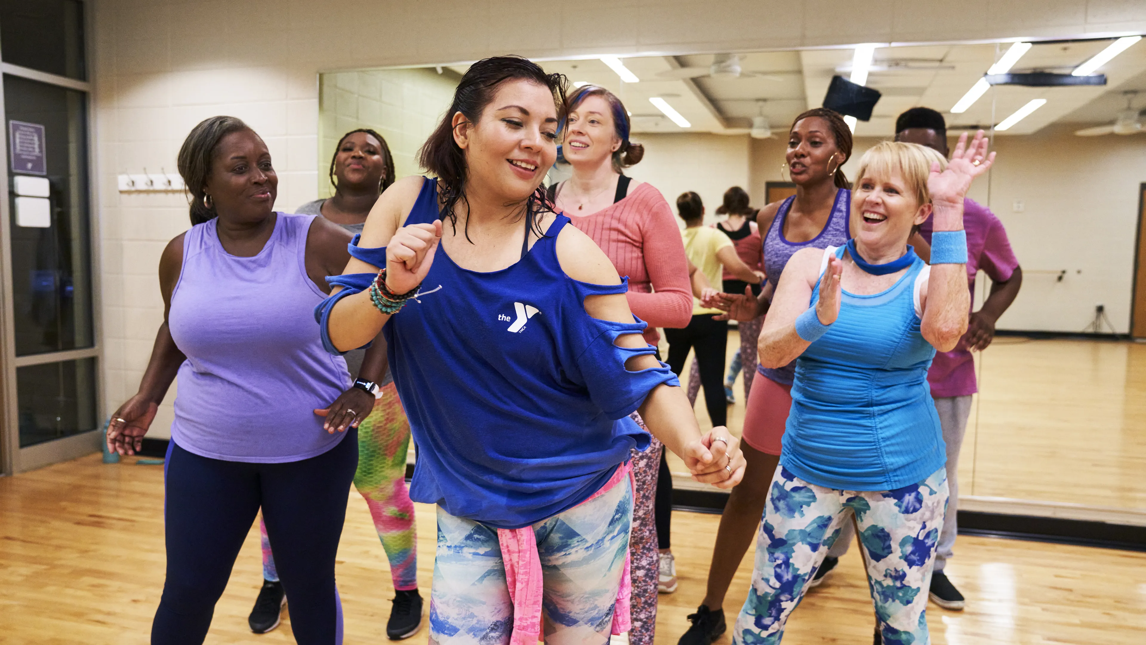 Group of people enjoying fitness class. Women and men dancing in brightly lit studio. One woman in blue top is clapping and smiling.
