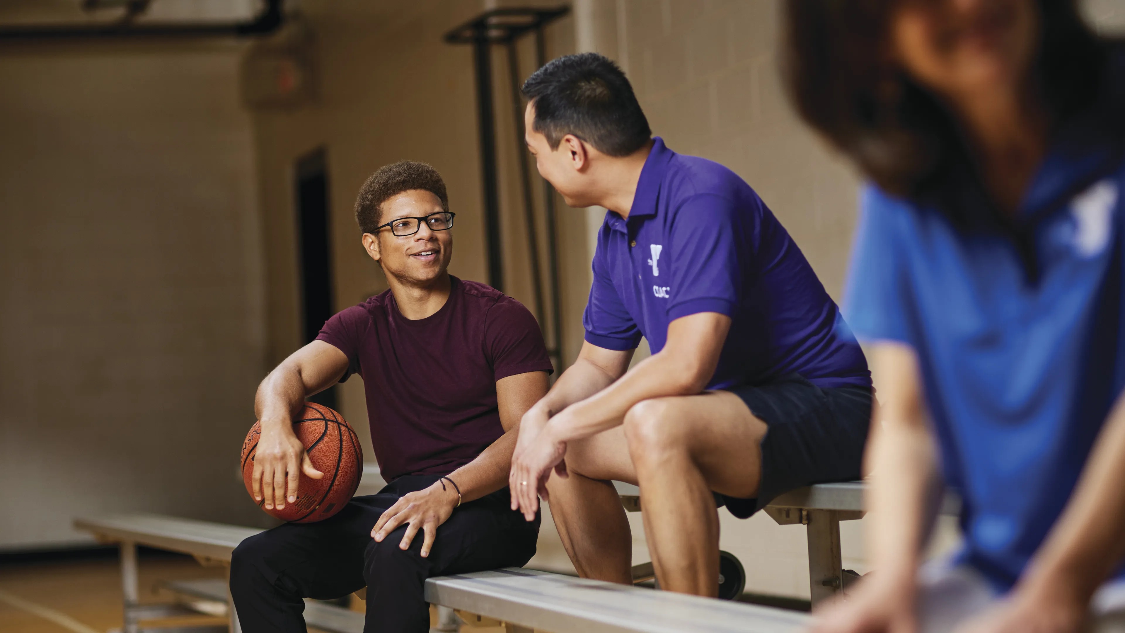 Two men and a blurred person sit on a bench in a gymnasium. One man holds a basketball. They appear to be talking during a break.