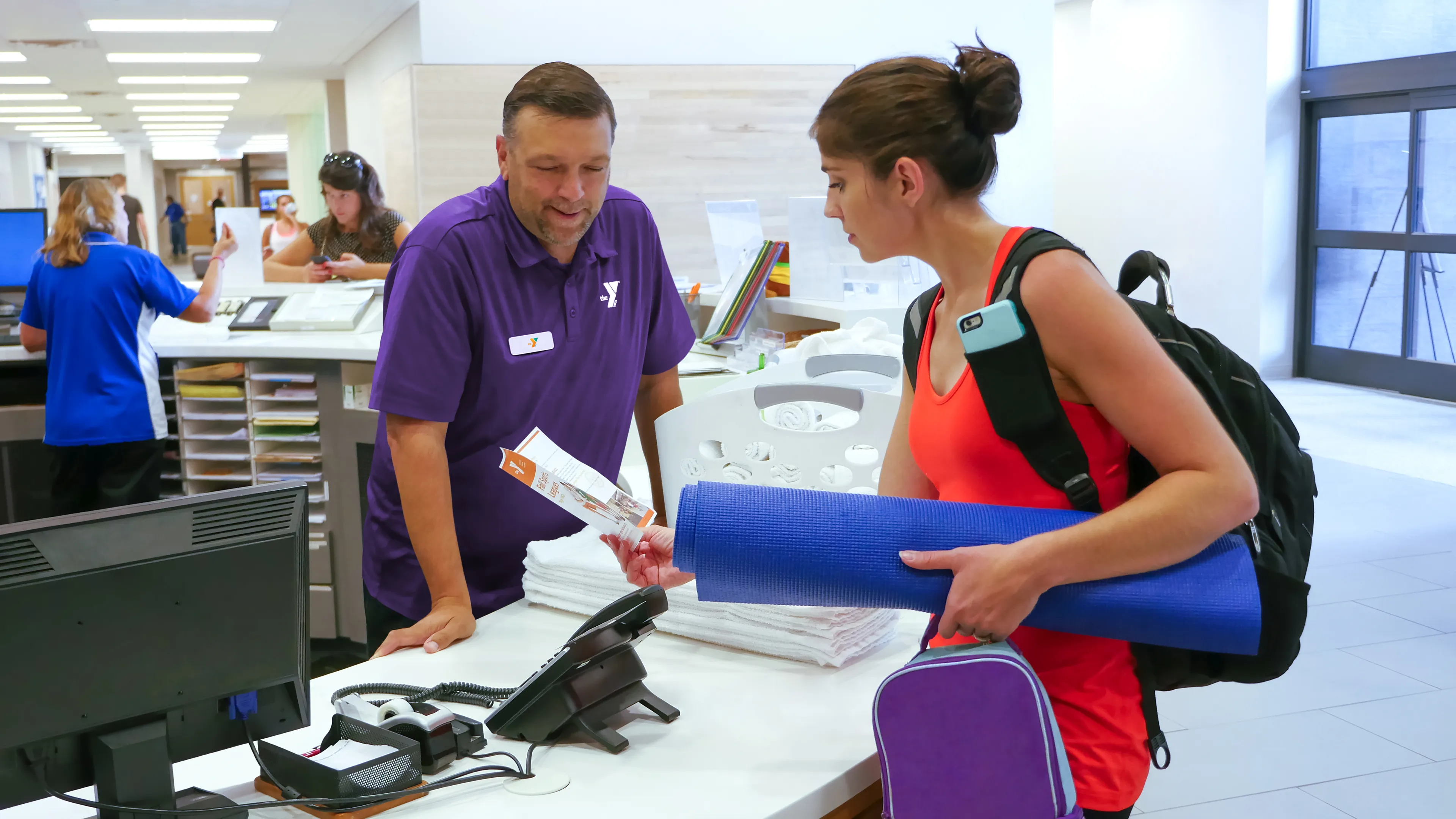 A woman with a backpack and yoga mat receives materials from a staff member behind the desk, possibly for class.