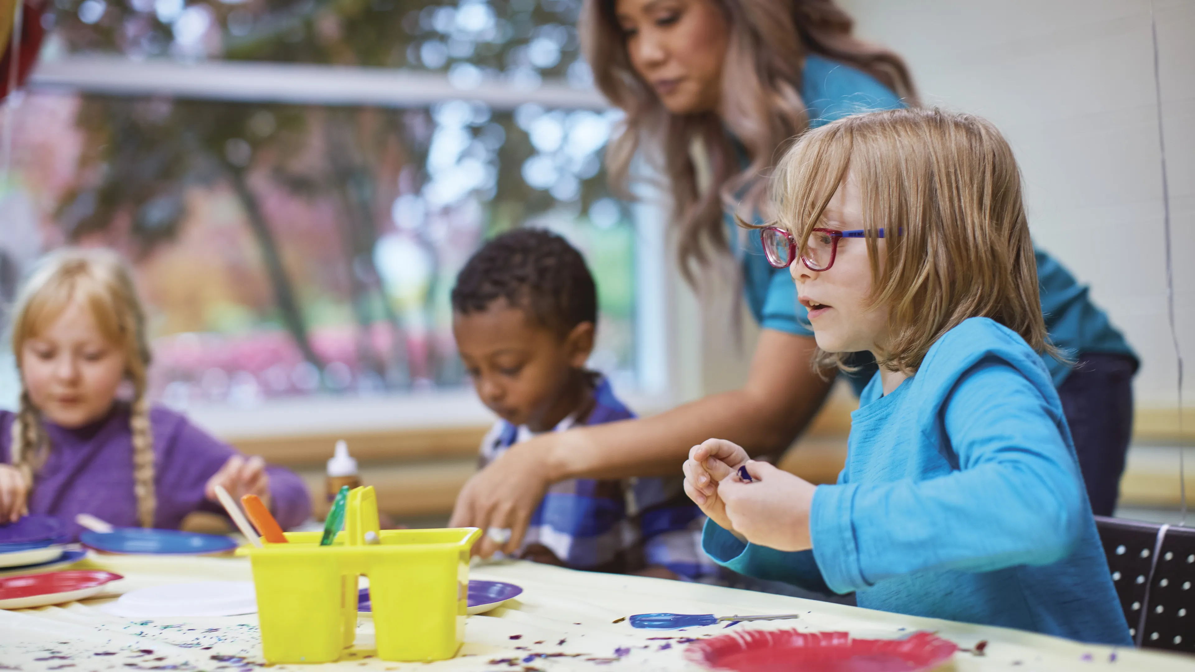 Children in childcare program gather around a table. An adult supervises as the kids work on crafts with paint and paper plates.