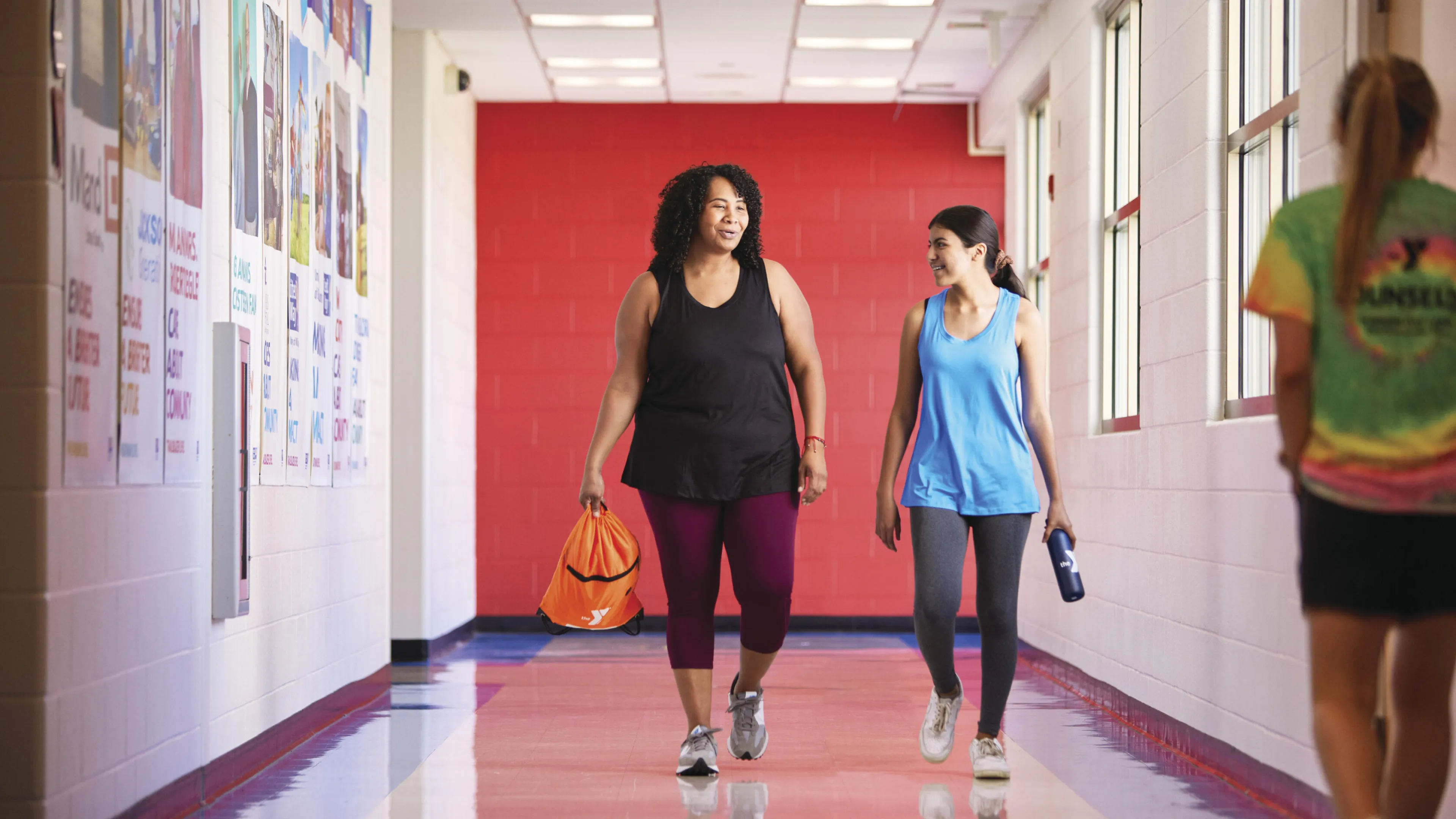 Two women stroll down a hallway, chatting; one carries an orange gym bag, the other a water bottle.  A third person, wearing a brightly colored shirt, walks ahead.  Colorful posters line the hallway walls.
