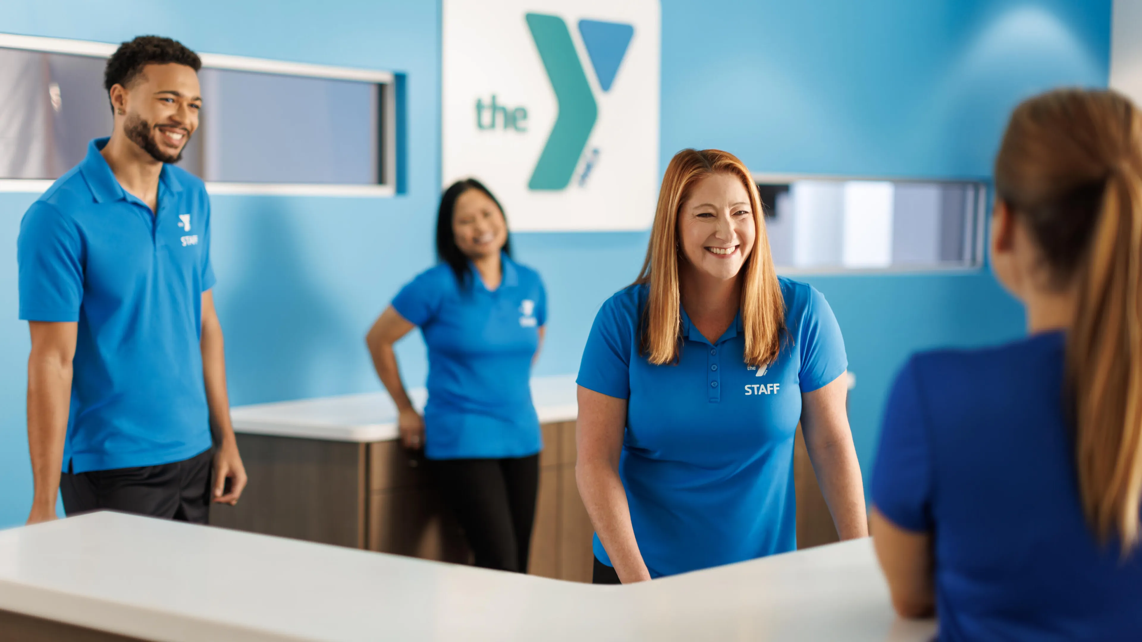 Smiling staff members at a reception desk greet a visitor.  One staff member's shirt says "STAFF".  Happy atmosphere.