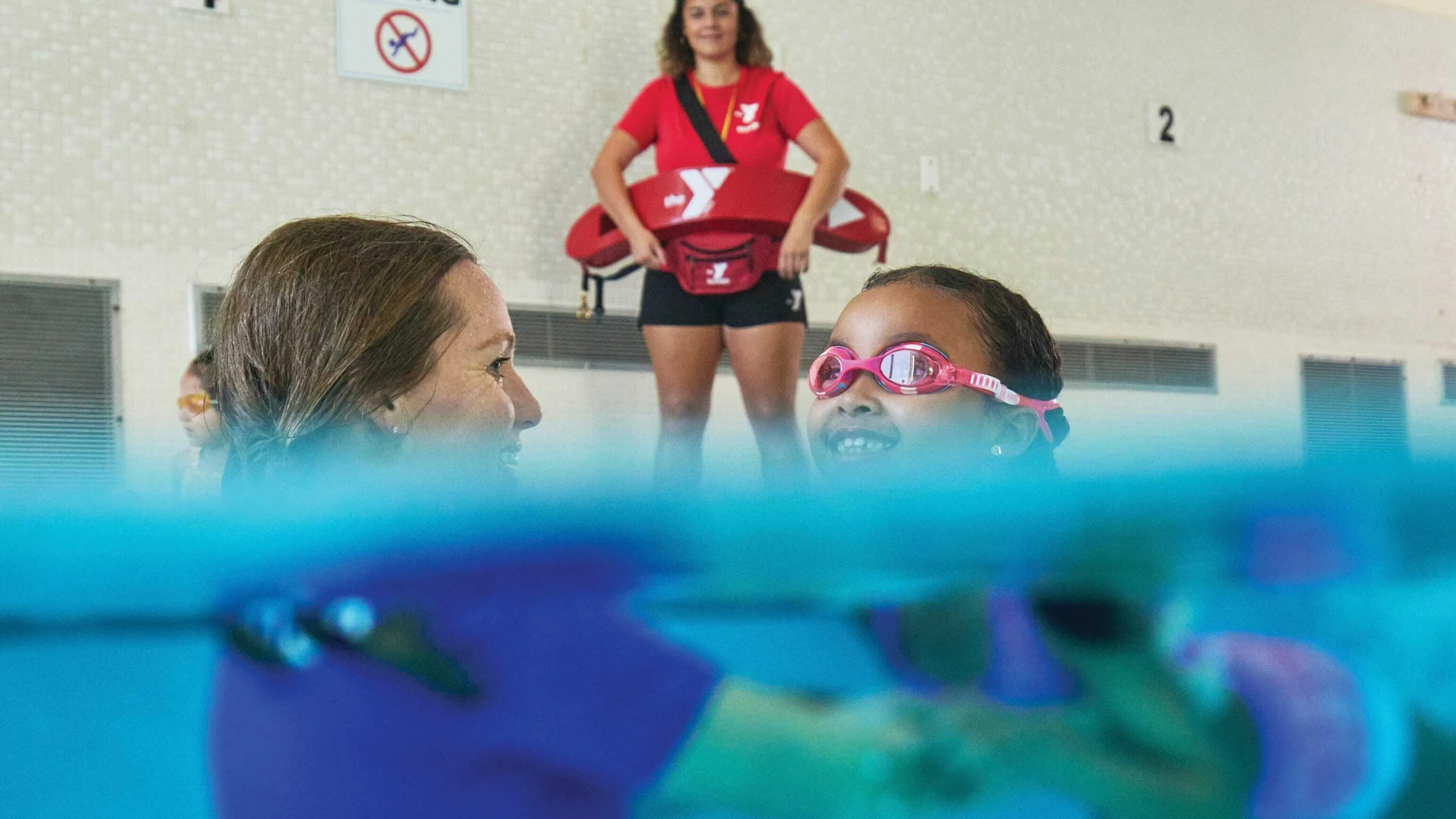 Smiling child in pink goggles learns to swim with an instructor; lifeguard watches over them at indoor pool, safety first.
