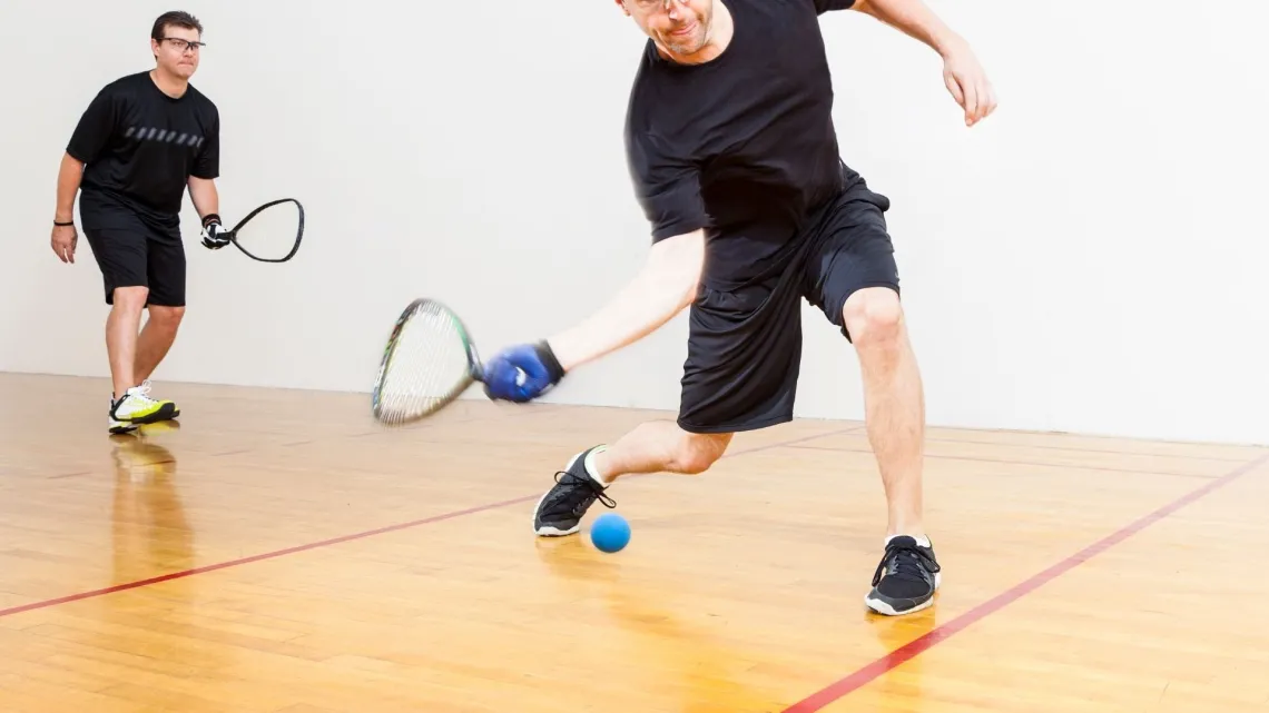 Two men playing racquetball in a court. One swings the racquet, preparing to hit a ball near the floor, other one is ready to receive.